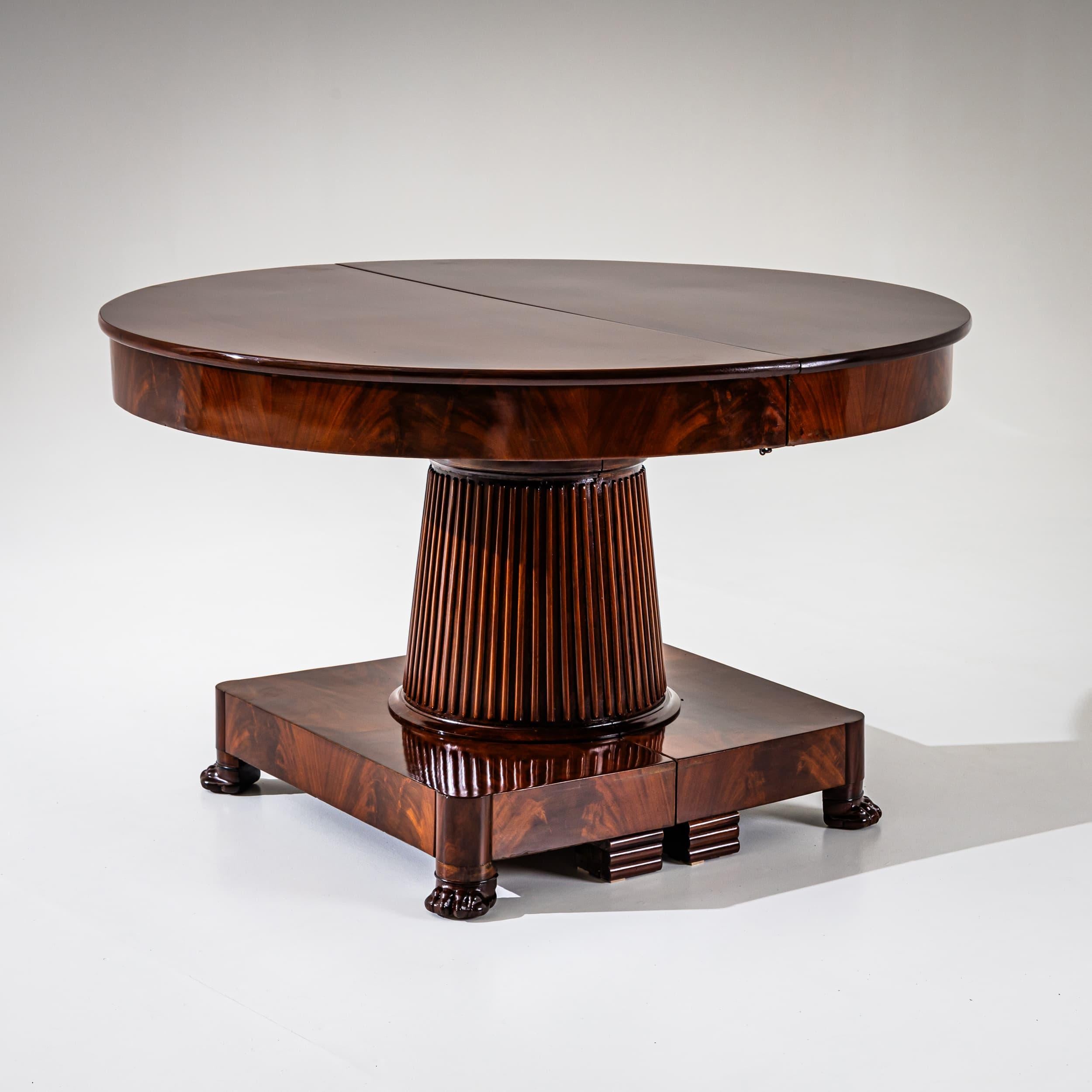 Elegant neoclassical extending table with a round mahogany top supported by a fluted, robust pedestal column. The base rests on a square platform with carved paw feet and profiled stabilizing plinths. The table can be extended in stages up to a