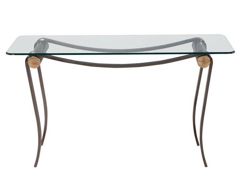 Neoclassical glass top chrome base console table. America, circa 1980’s. Curved metal frame with accented brass plated front details. Completed with a thick rounded glass top. Glass is original and has minor scratches consistent with age and use.