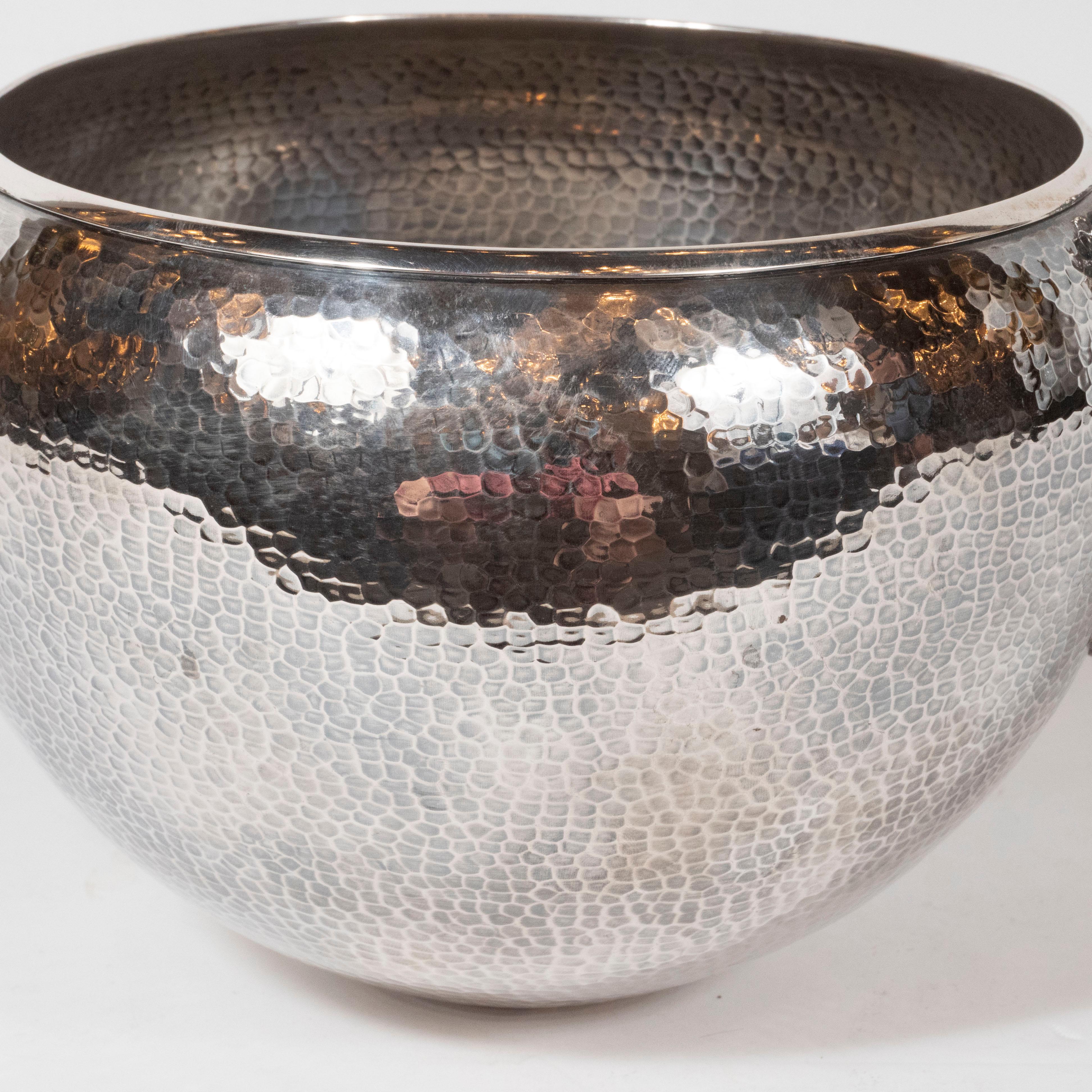 This sophisticated modernist bowl was realized in the United States. It features a spherical body with a circular mouth; a hand hammered dappled texture throughout; and two figurative male handles, inspired by Greek classicism that protrude from the