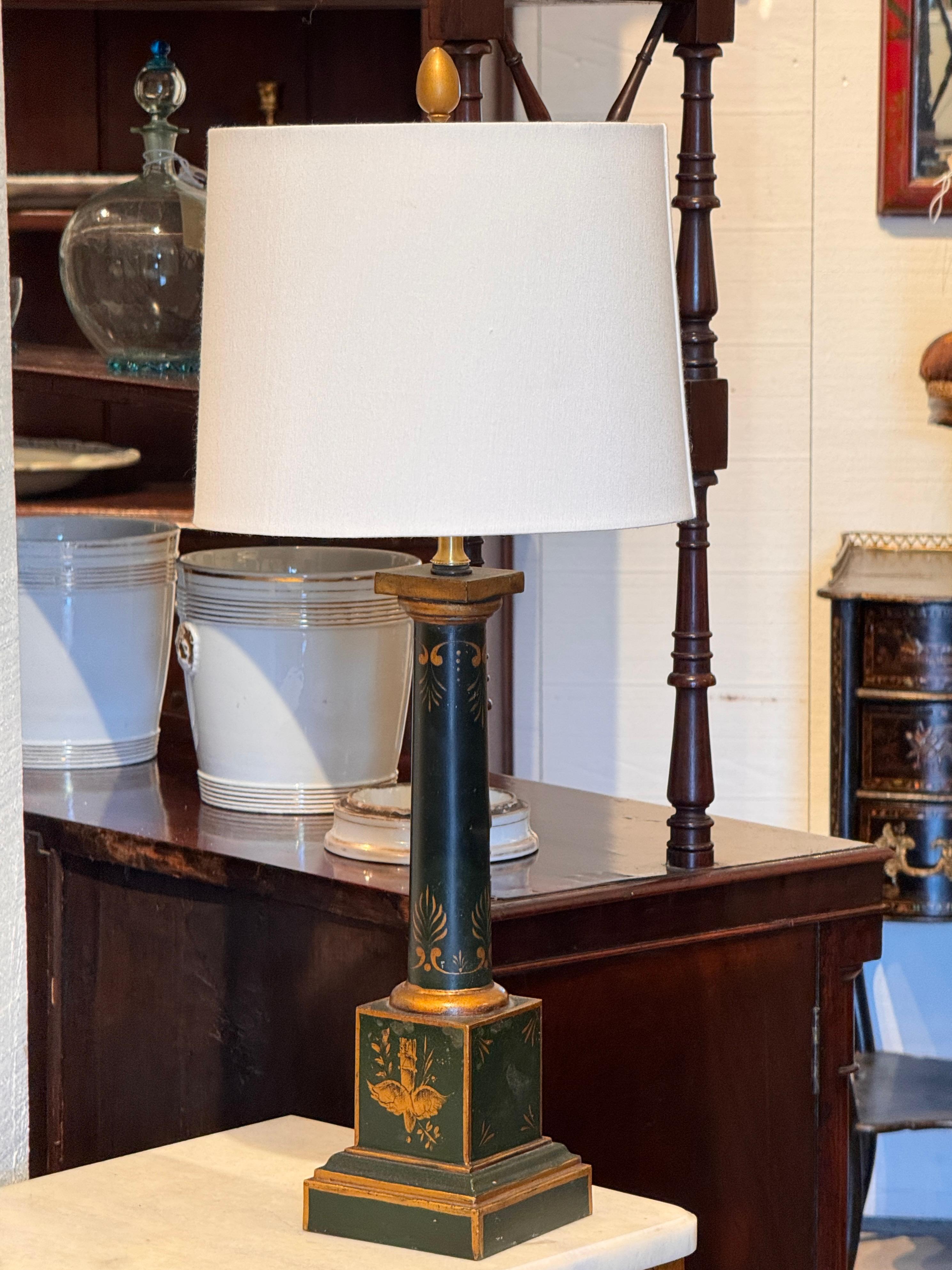 Painted in a deep green with gilt neoclassical motifs, this tole lamp features a columnar form on a stepped square base. The hand-painted decoration includes stylized foliate elements and classical emblems on each side of the plinth, contributing to