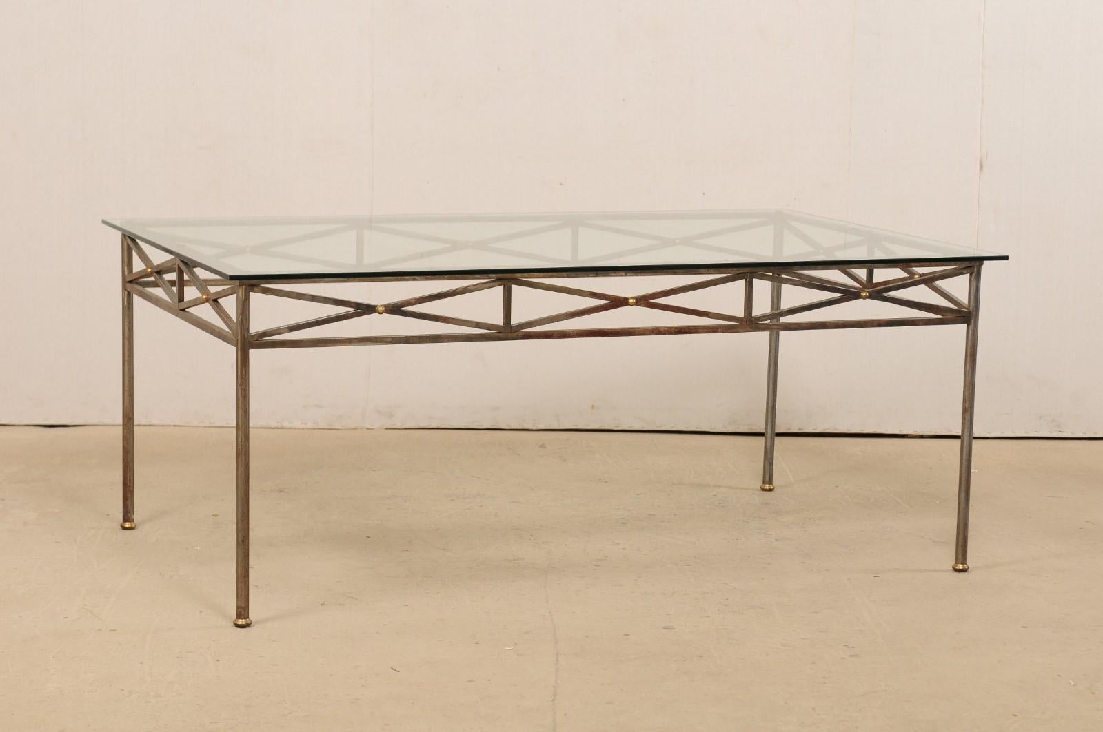 An American-made rectangular-shaped glass top table from the late 20th century. This vintage table has a neoclassical inspired design, with metal skirt comprised of an open, X-shaped pattern with petite brass ball accents at each crossing center.