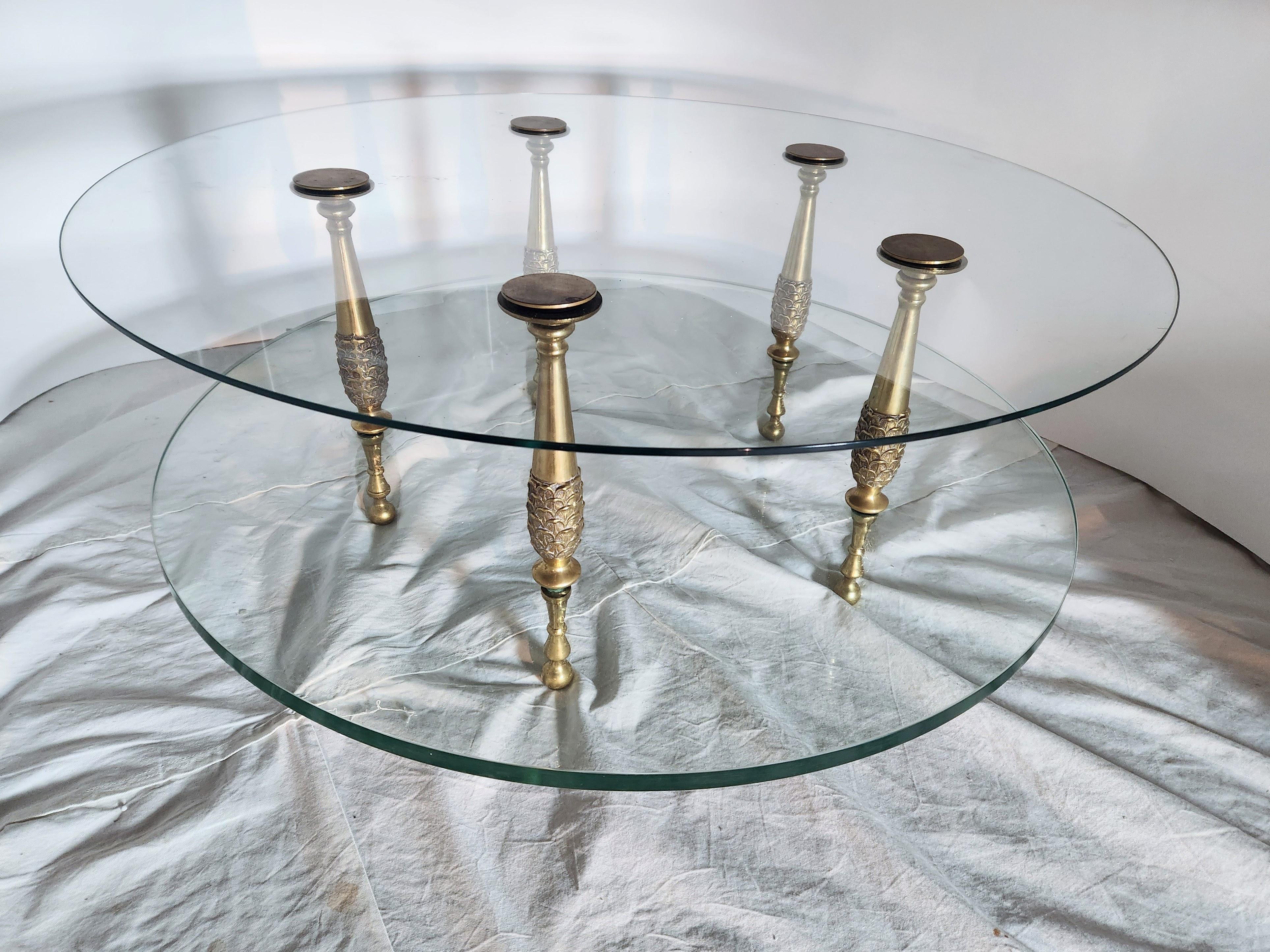 Insolite table basse néoclassique italienne à deux niveaux en verre, datant des années 1960, en bronze coulé.
Cinq supports segmentés en bronze allongé en forme de pomme de pin soutiennent les deux étagères en verre qui se terminent par des disques