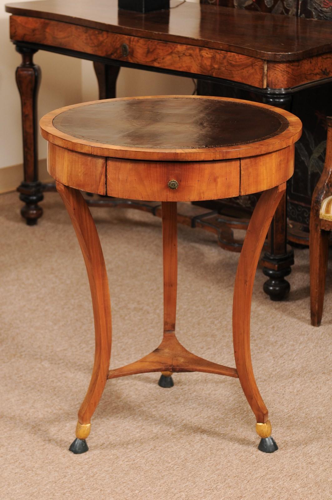 The neoclassical Italian gueridon in fruitwood with circular embossed brown leather top, one drawer and supported by three concave legs with lower shelf and hoofed feet. 

 