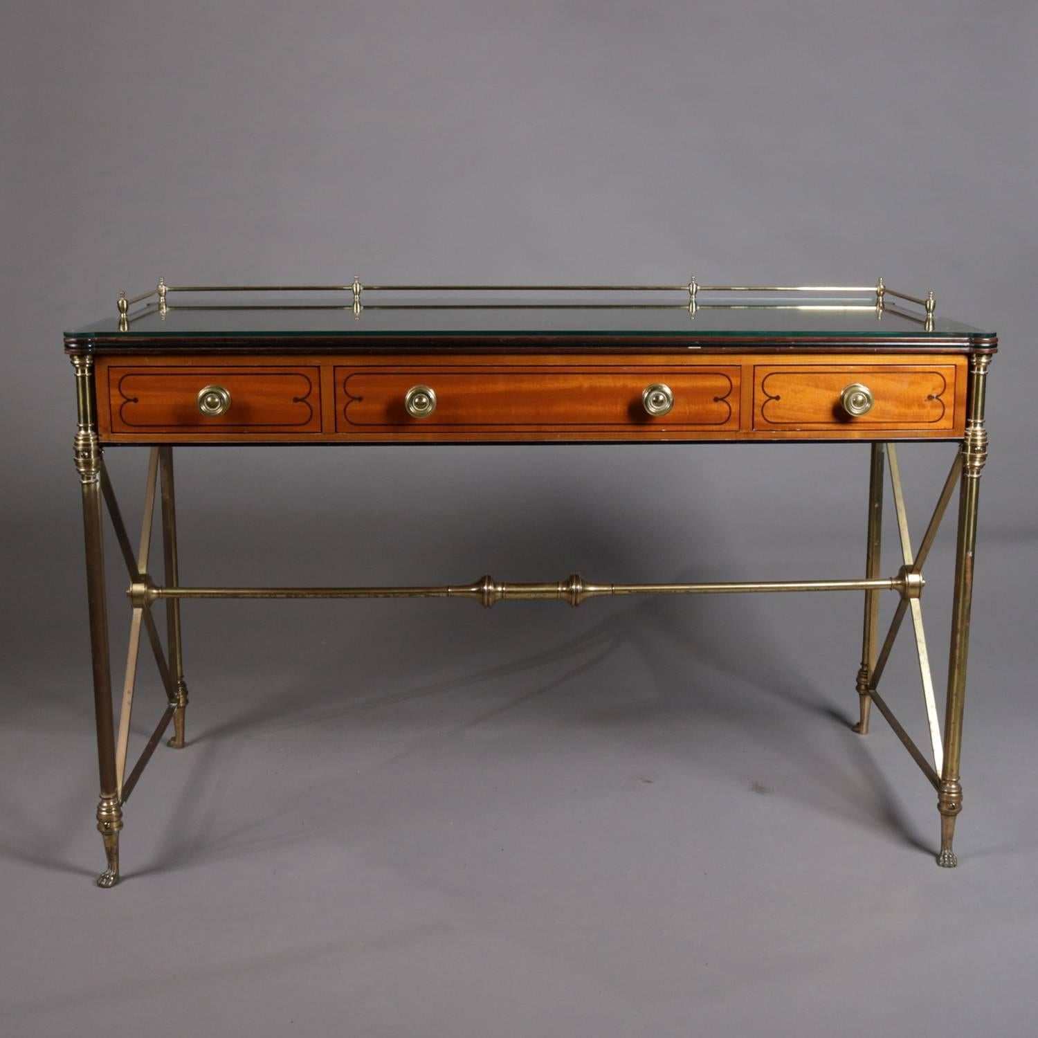 Neoclassical writing desk by Kittinger features three-drawer satinwood case with shaped top having brass gallery and glass covering, seated in brass frame with X-form support with floral medallion and between column-form legs terminating in paw