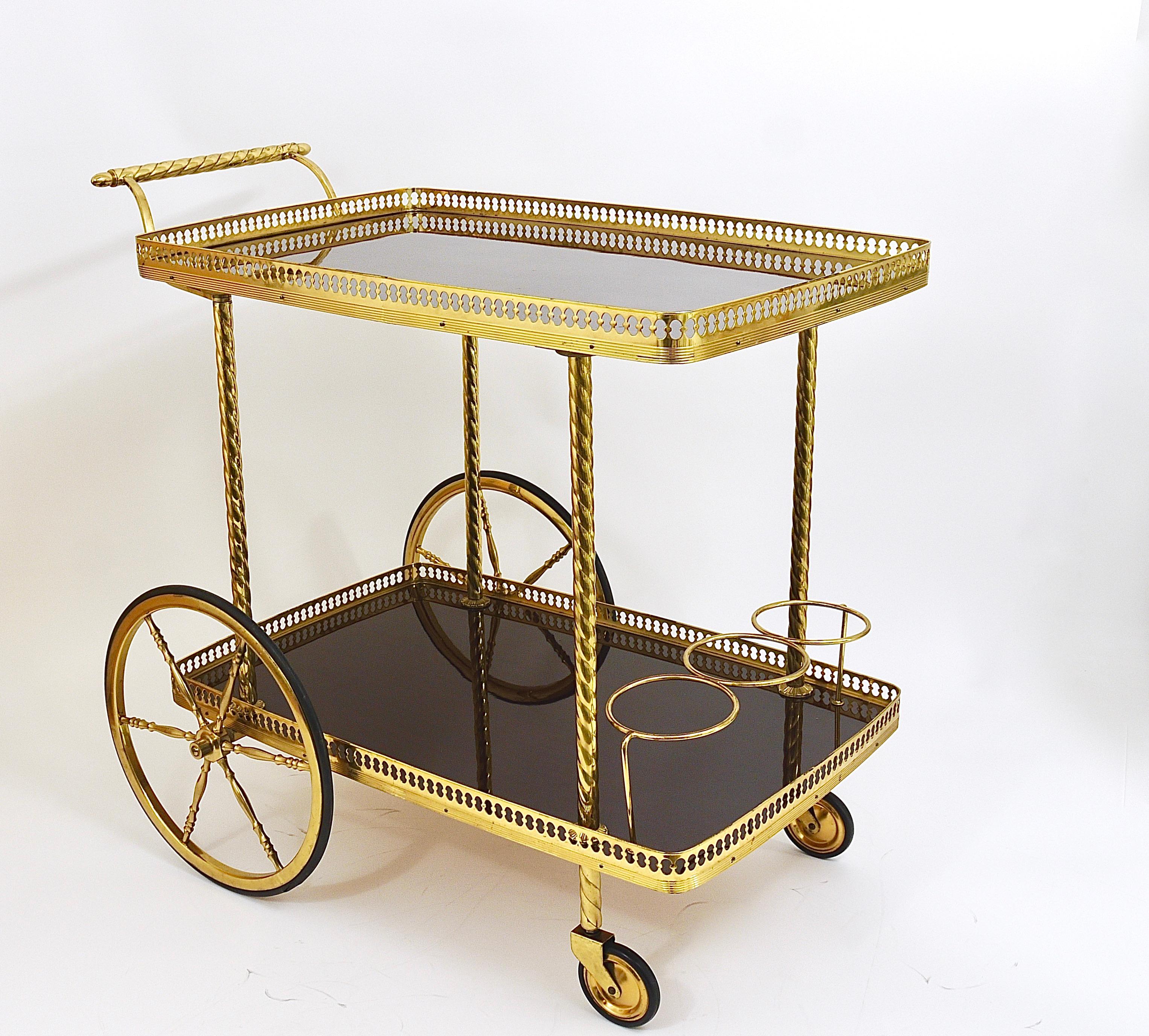 An exquisite and elegant French Hollywood Regency brass bar cart/ drinks serving trolley from the 1950s. A beautiful Midcentury piece of furniture with two glossy mahogany plateaus, decorated by pierced brass galleries. Standing on four lovely