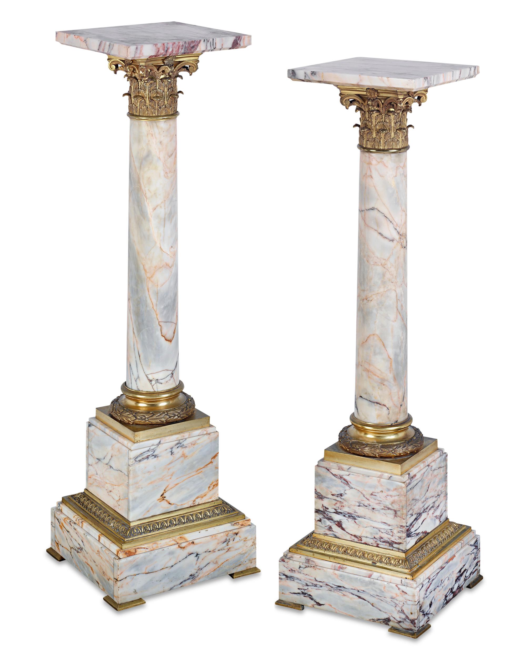 A classic Neoclassical style sets apart this stately pair of colorfully-veined white marble pedestals. Taking the form of Corinthian columns with brilliant gilt bronze capitals, the impressive pieces can serve as display pedestals or as stunning