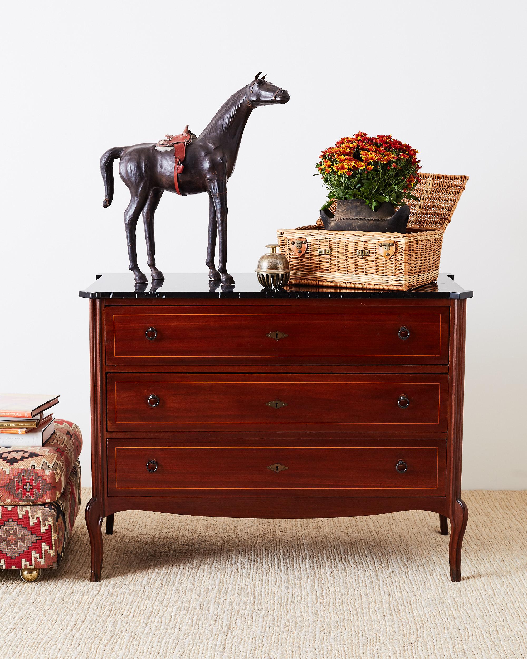 Stately mahogany neoclassical style commode or chest of drawers featuring a polished black marble top. The mahogany case is fronted by three legs drawers with satinwood thread inlay and brass ring pulls. Each corner has a reeded column design with