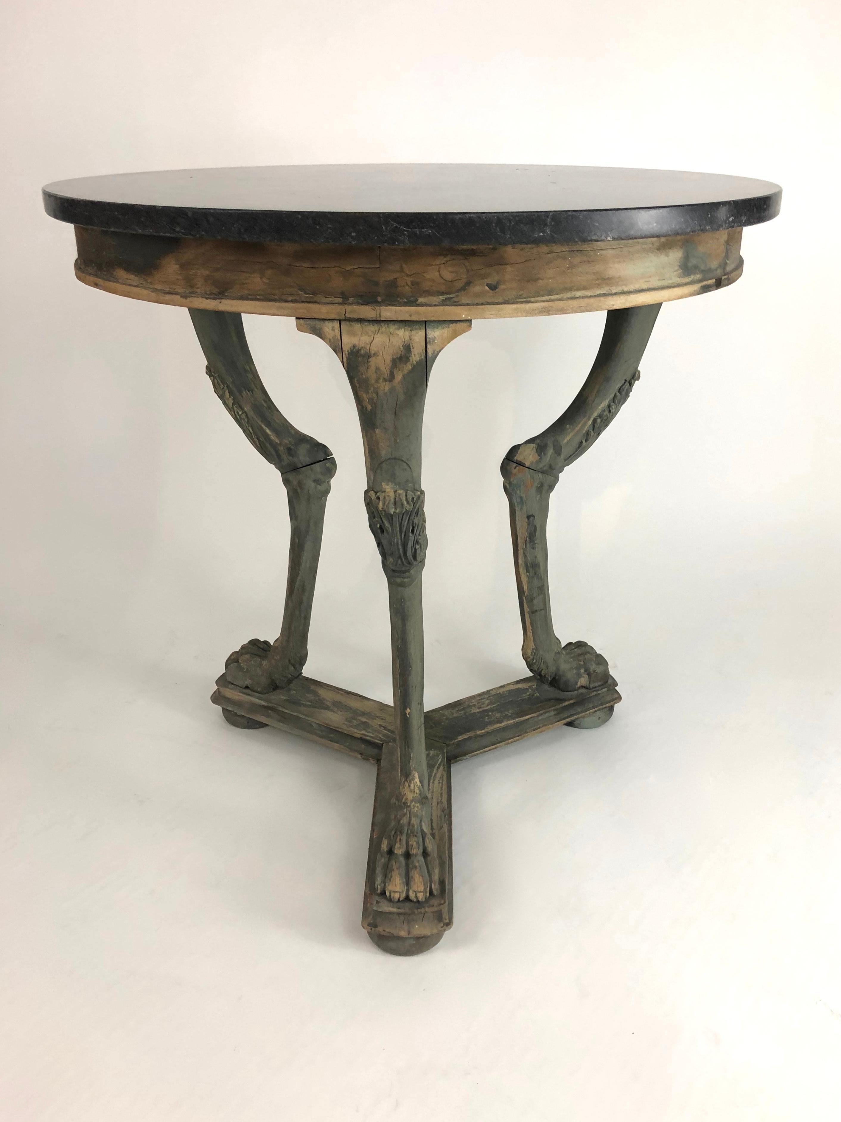 A neoclassical gueridon side table with charcoal grey marble top supported by a carved wood tripod base with acanthus leaf carved legs and animal paw feet joined by a three way plinth raised on bun feet, in well worn old grey painted finish. This