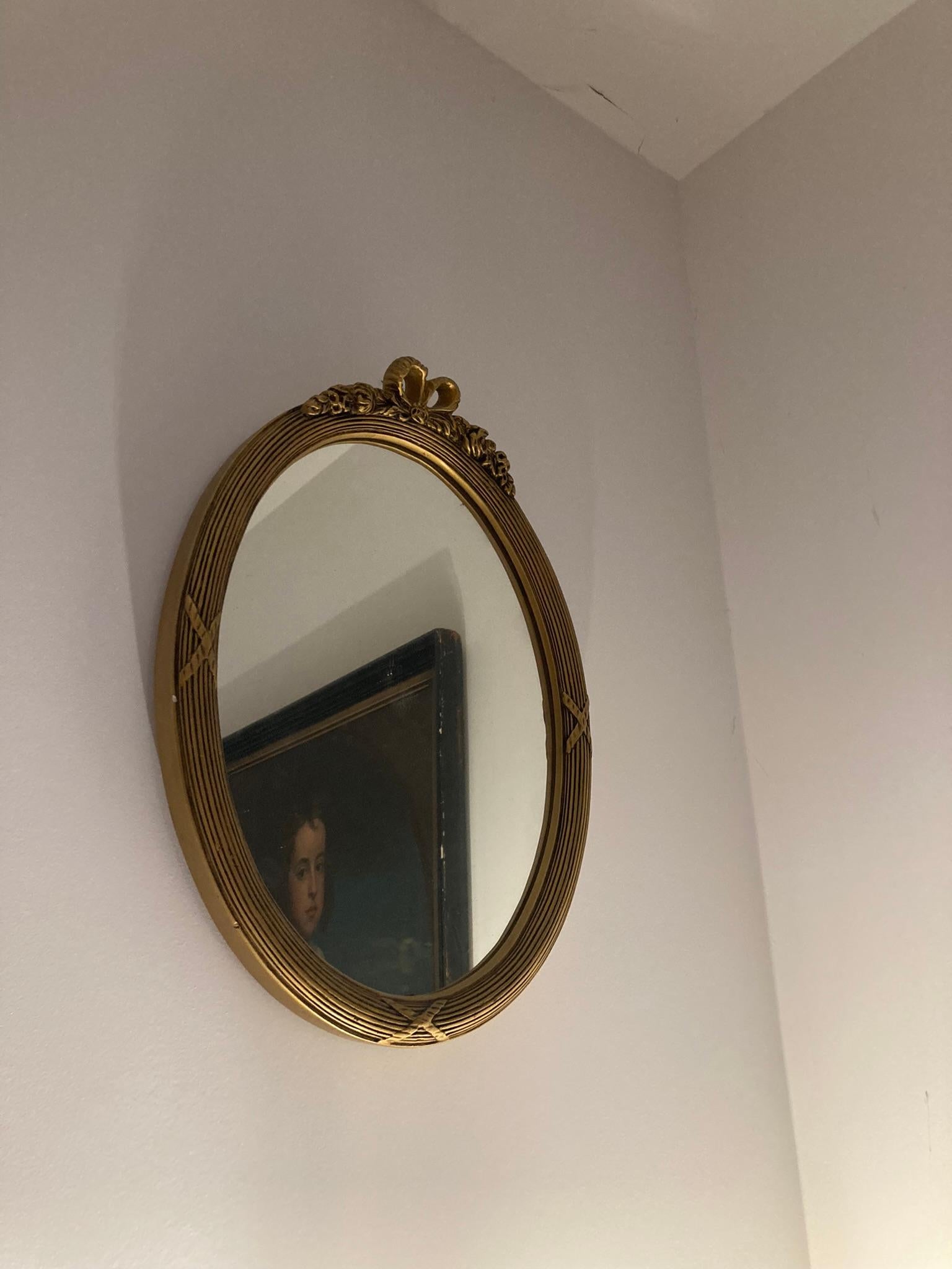 Espejo de pared ovalado de madera dorada de estilo neoclásico, de principios del siglo XX. El marco es de madera y está coronado con una cinta en la parte superior. El cristal del espejo presenta un borde facetado. La parte trasera del espejo se ha
