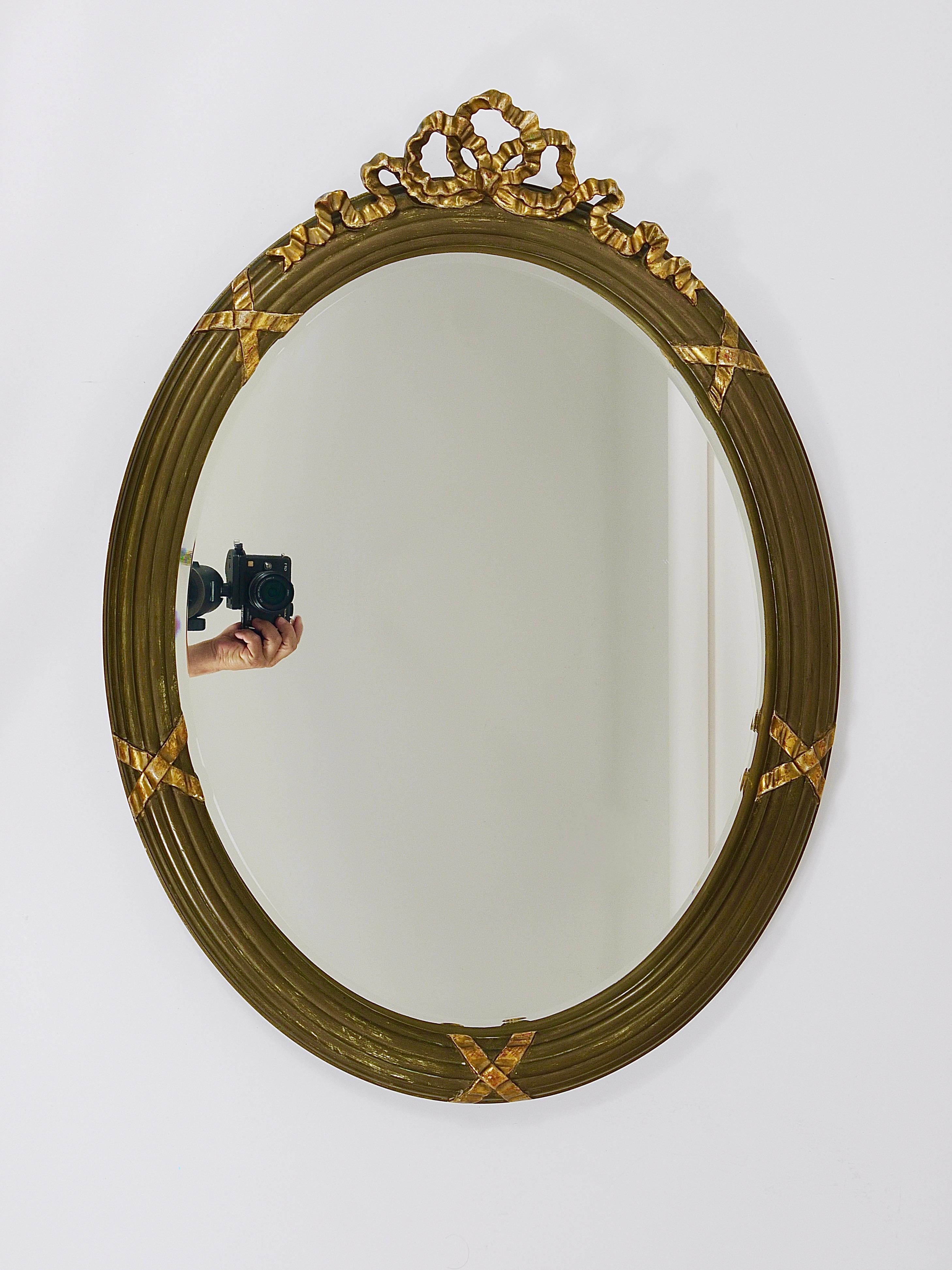 A beautiful and decorative oval gilt wood wall mirror in the Neoclassical style, dating from the early 1900s. The frame is made of wood, partially finished with hand-applied antique gold leaf and crowned with a charming, hand-carved and detailed