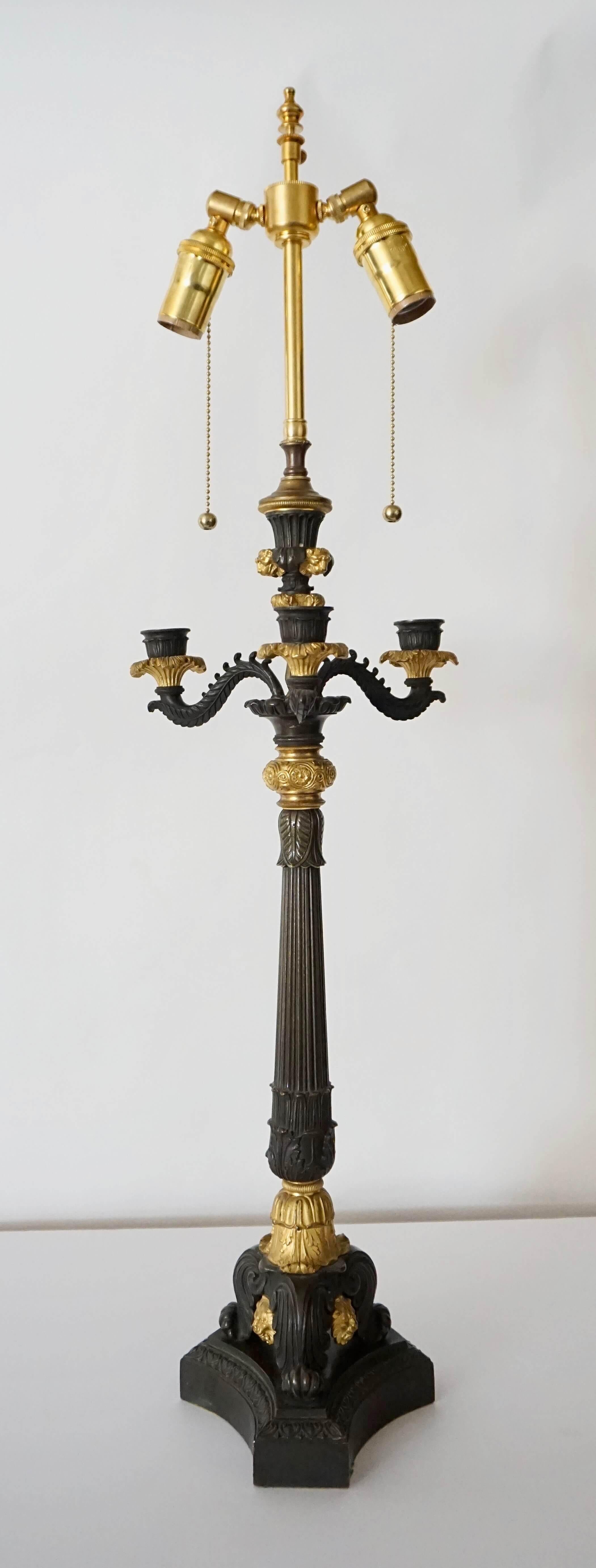 A circa 1825 neoclassical English Regency candelabra converted to electricity having solid patinated and ormolu bronze body with acanthus, foliate, lambs tongue, and lions head motifs; the three scrolling candle arms atop reeded shaft on tripart