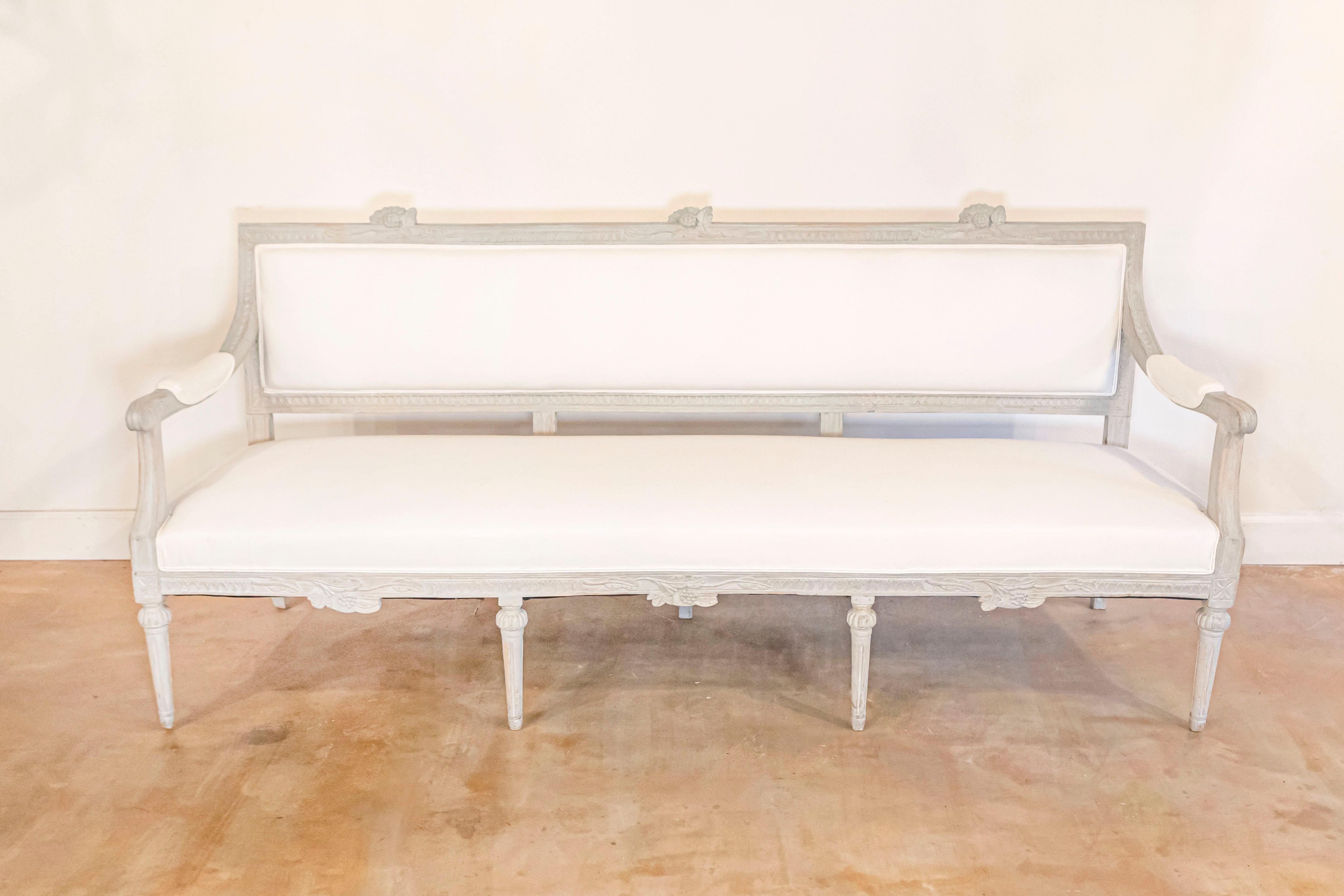 A Neoclassical Revival Swedish carved and painted bench from the late 19th century with scrolled open arms, fluted legs, floral motifs and new upholstery. This Swedish bench features an upright rectangular back that has been reupholstered in a