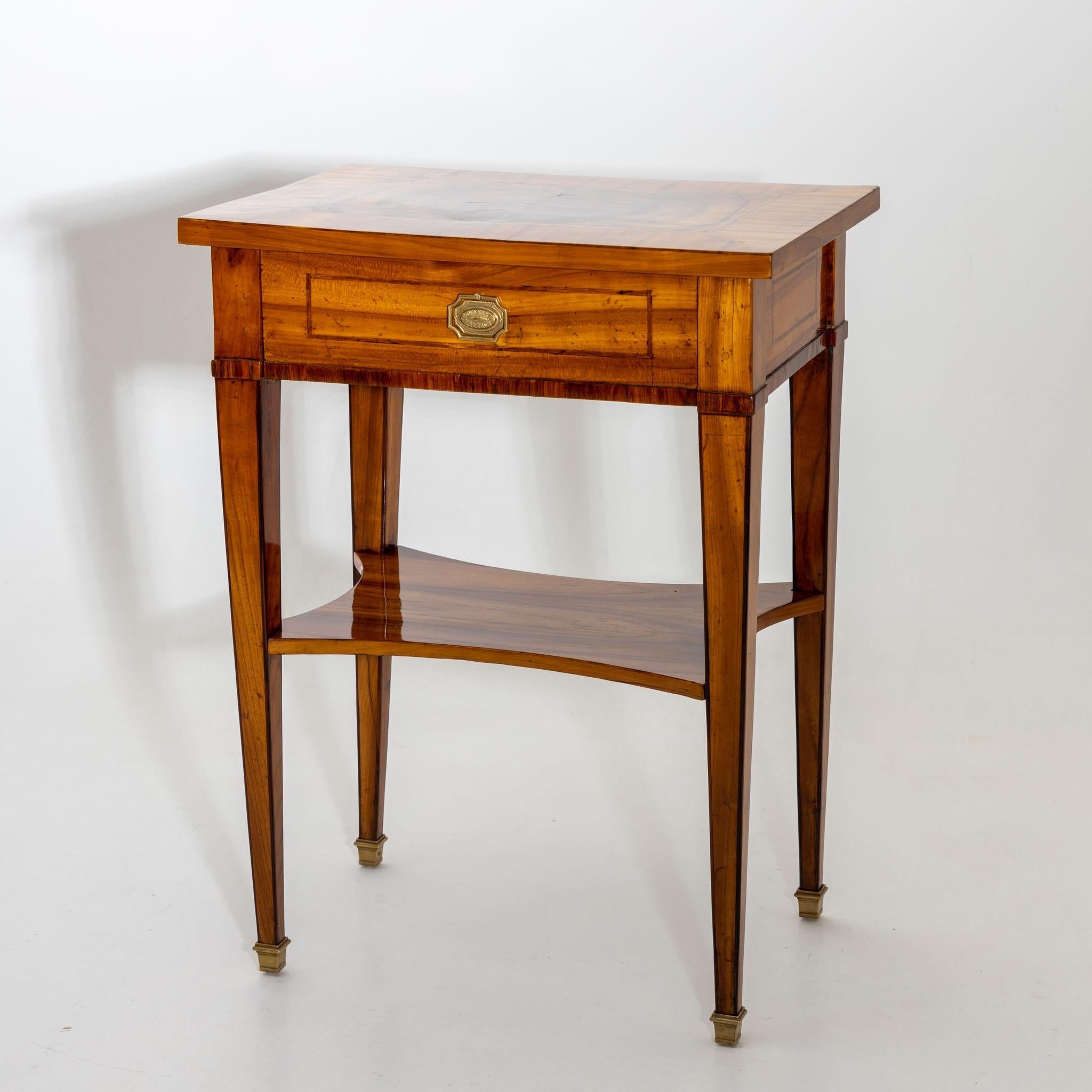 Small side table on high square pointed legs with brass sabots and a drawer including a clipboard. The drawer is divided into smaller compartments. The table is veneered in cherry and hand polished.
