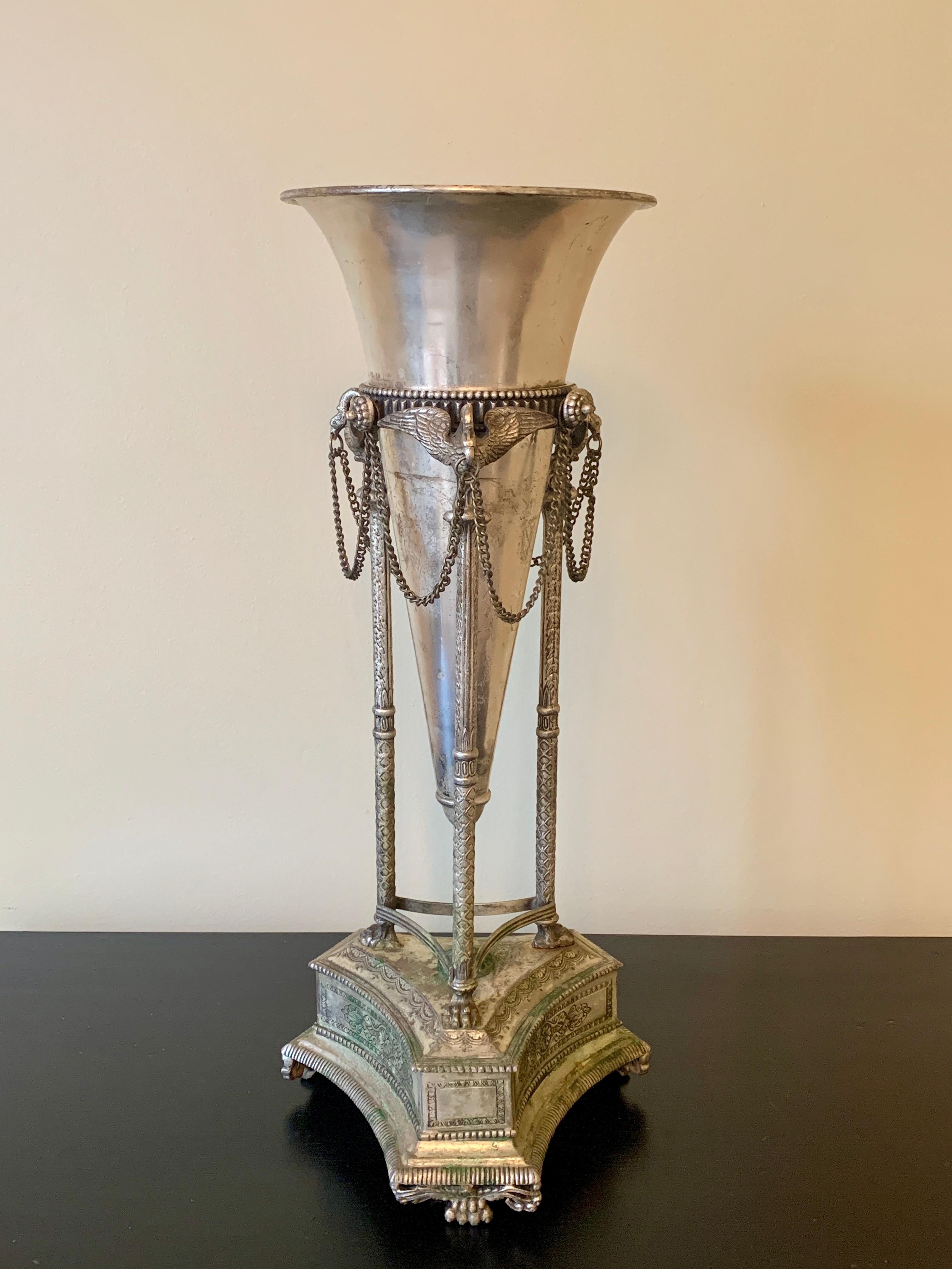 A stunning vintage silver plated neoclassical style vase. The stand rests on three paw feet, a stylized base, another set of three paw feet which support three stylized columns, culminating in a trio of swans holding a swagged metal chain in their