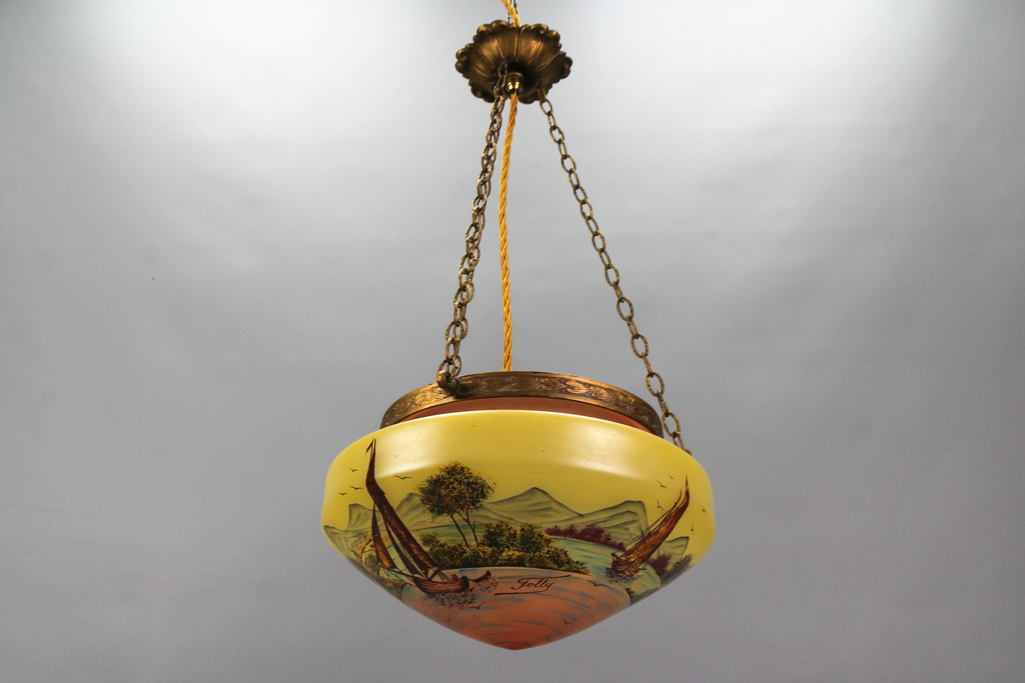 Neoclassical style brass and glass pendant light with hand-painted landscape from circa the 1920s.
This stunning Neoclassical-style pendant light fixture features a yellow and pale salmon color glass lampshade - an oval bowl. The lampshade is
