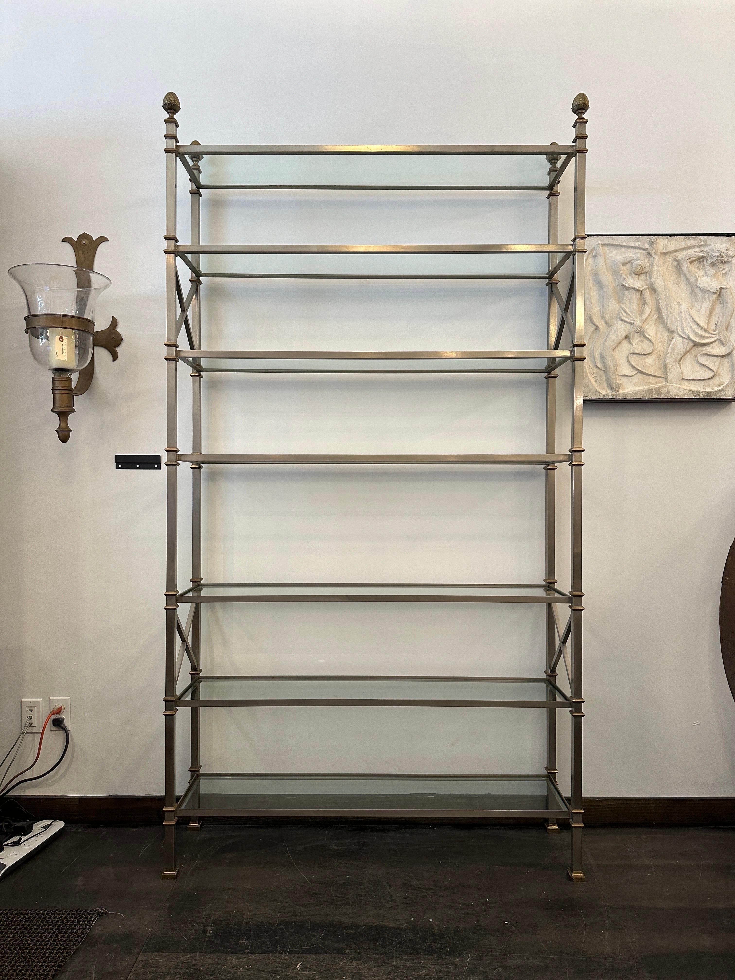 Maison Jansen, un'étagère con 7 ripiani della metà del XX secolo.  Telaio in acciaio spazzolato con decorazioni a ghianda in ottone e barelle con design a X.  Sette spessi ripiani in vetro possono contenere oggetti molto pesanti e il telaio quadrato