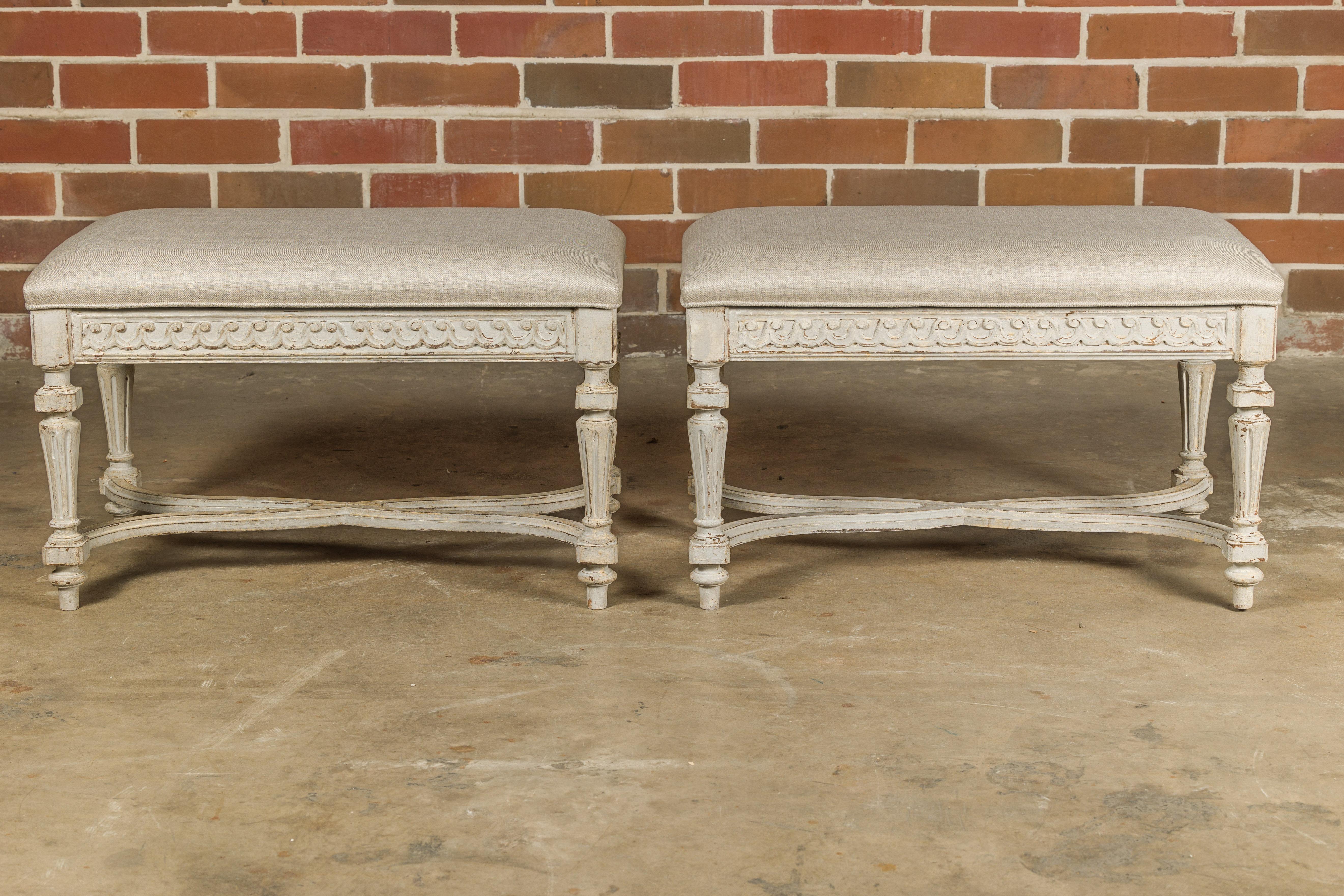 A pair of French Neoclassical style painted wood benches from the mid 20th century, with carved Vitruvian scroll frieze, fluted legs, X-Form cross stretchers and custom upholstery. This pair of French Neoclassical style painted wood benches, crafted