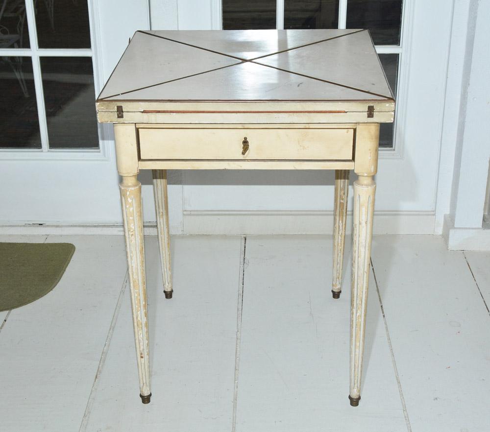French envelope game table. By opening the four hinged triangular sections the small end table in the neoclassical style becomes a square game table with a blue inset panel. Painted cream, the table has gilt trim, fluted legs with brass tips and a
