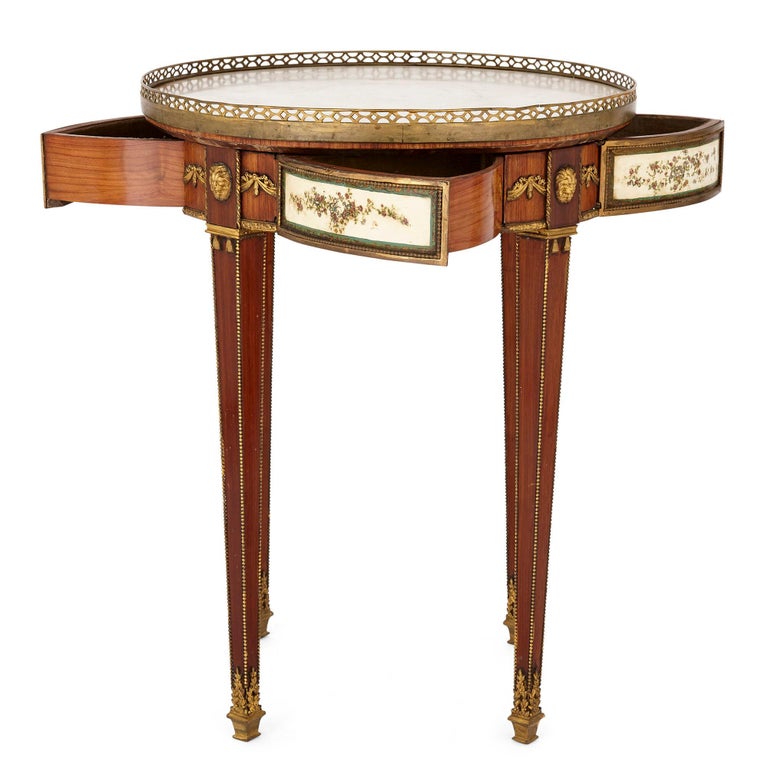 This Neoclassical style mahogany side table is inset with a circular white veined marble top that is bounded by a gilt bronze gallery. Below the top, set into the apron, are four pivoting drawers, the fronts painted above white ground with floral