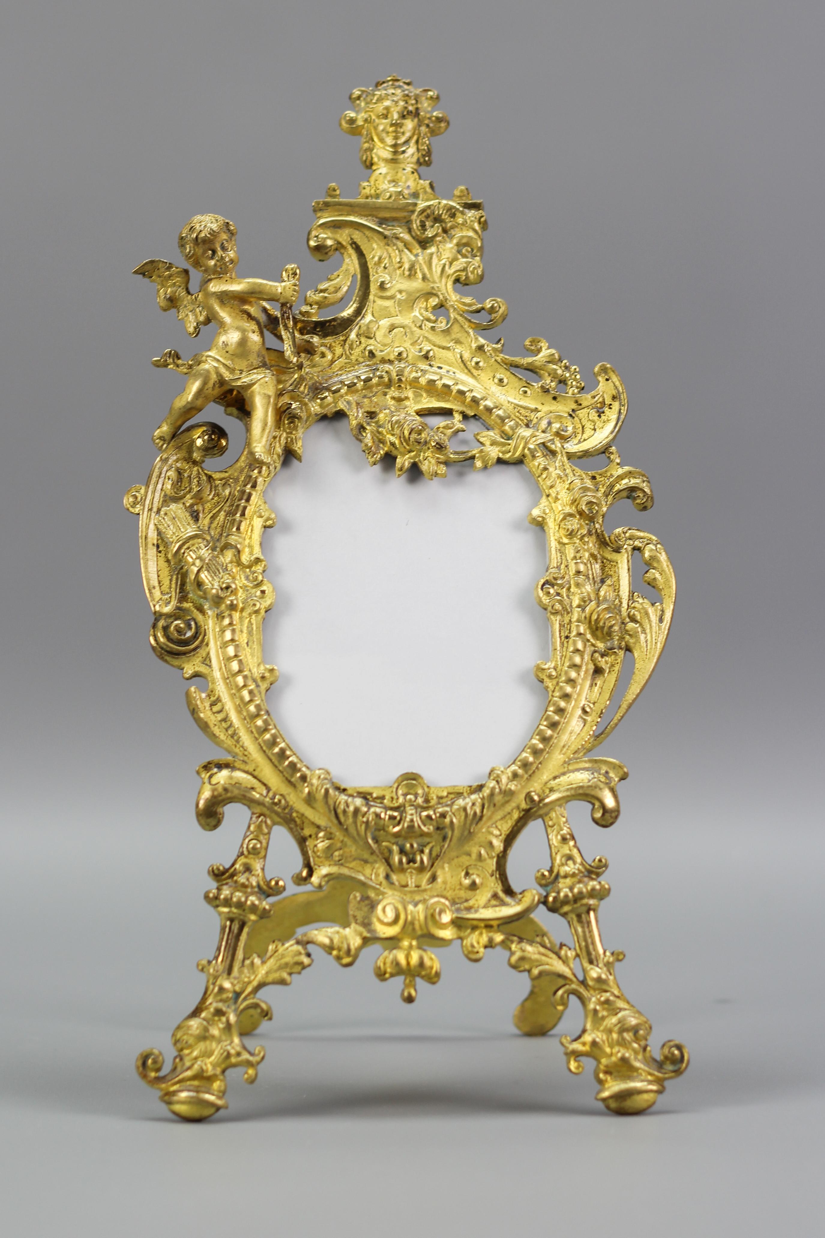 An adorable gilt bronze photo frame, richly decorated with a cherub and Neoclassical or Louis XVI style motifs like a face of a goddess, garlands of flowers, foliage, darts, and a face of a satyr. This beautiful oval-shaped picture frame features an