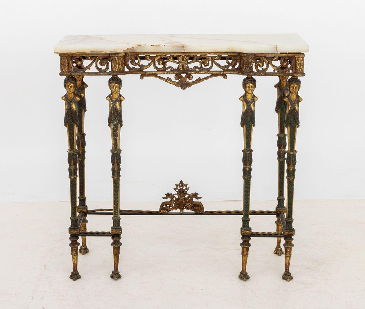 Mesa consola de metal dorado y ónice de estilo neoclásico, principios del siglo XX, con volutas caladas y motivos florales sobre seis patas cariátides y acanaladas unidas por camillas con acentos de forma retorcida sobre seis patas de garra. Alto