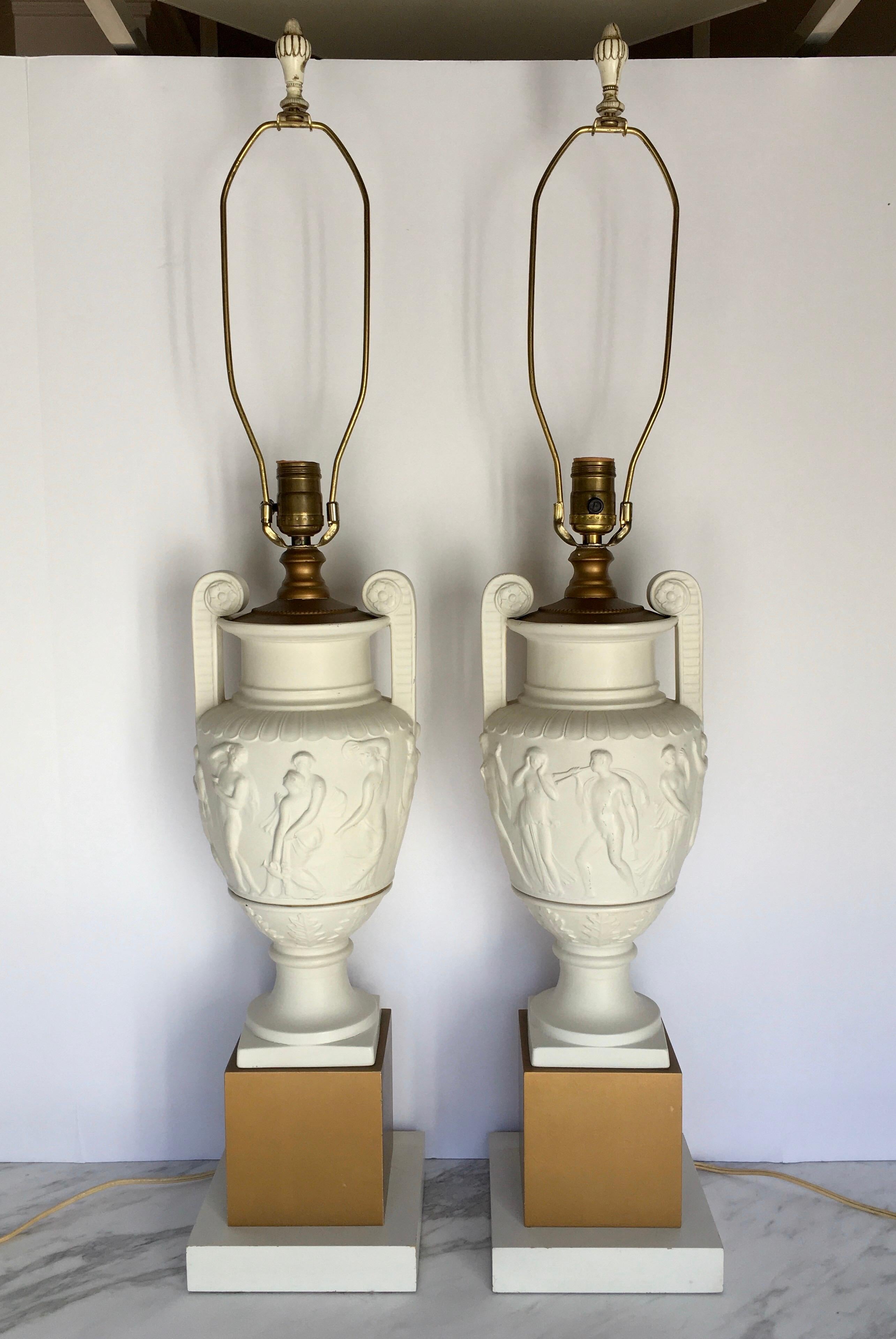 Pair of neoclassical style ceramic and wood urn table lamps mounted on square plinth gold wood bases. Plaster-like matte cream handled urns feature sculptural relief figures. Lamp shades not included. 

Measures: Height to harp: 35.5