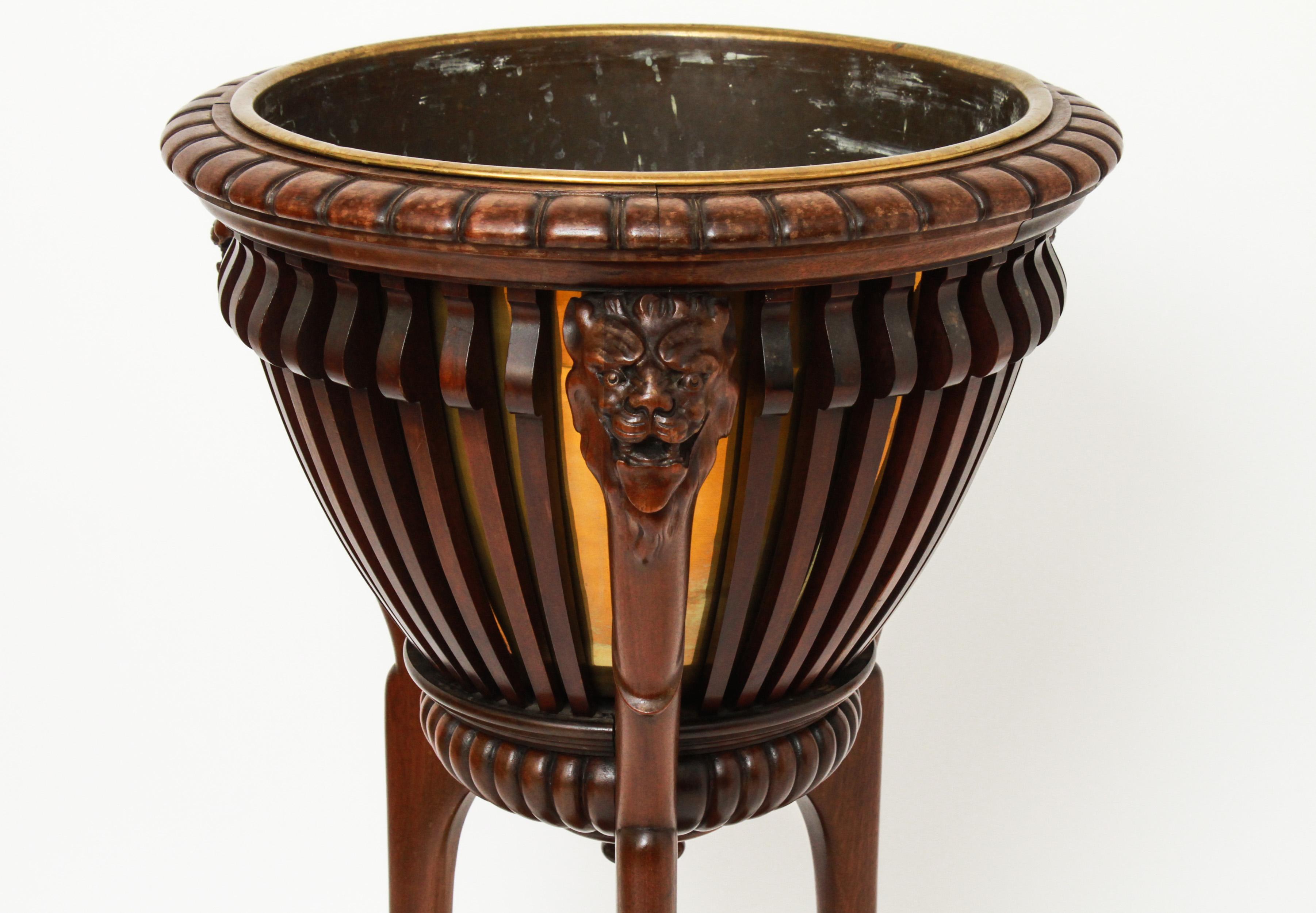 Neoclassical style hardwood jardinière with lion head motif. The piece has a carved top with snarling lion mask supports, interior fitted with a metal liner and rising on three cabriole legs conjoined by a lower shelf and terminating in paw feet. In