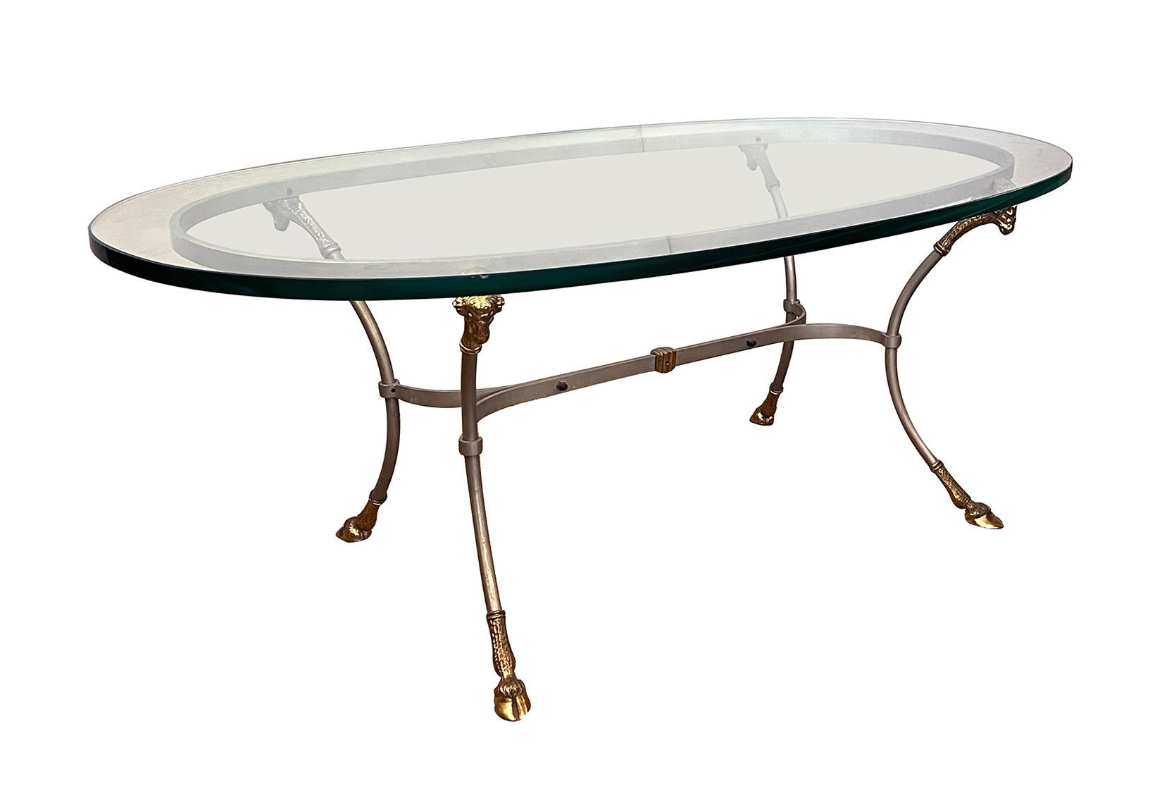 à la manière de la Maison Jansen, l'épais plateau de verre ovale reposant sur un corps en acier conforme, avec quatre supports élégamment évasés, coiffés de têtes de béliers en laiton et terminés par des pieds en sabot ; le tout relié par des