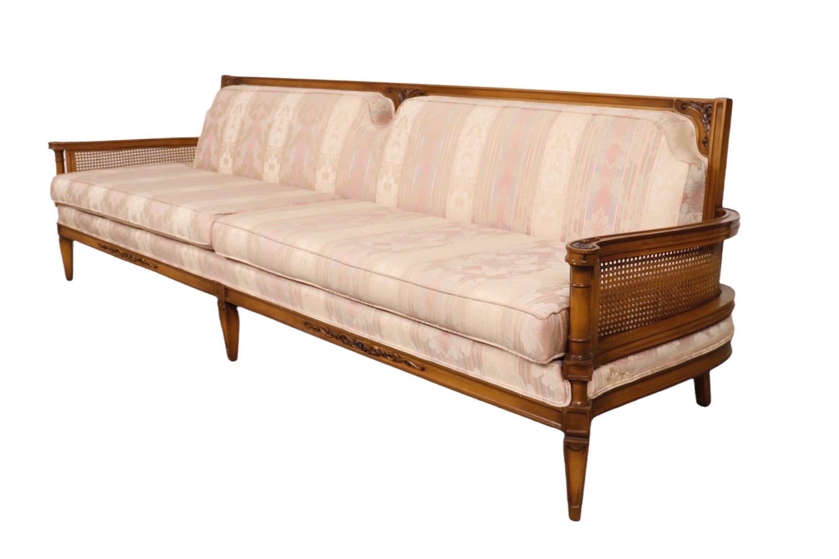 A large neoclassical style caned sofa. Clean lines throughout with a flat crest rail, carved at the center with scrolled acanthus. Outward rolled arms are topped with rosettes above caned side panels. Upholstered with a tight seat back and two long