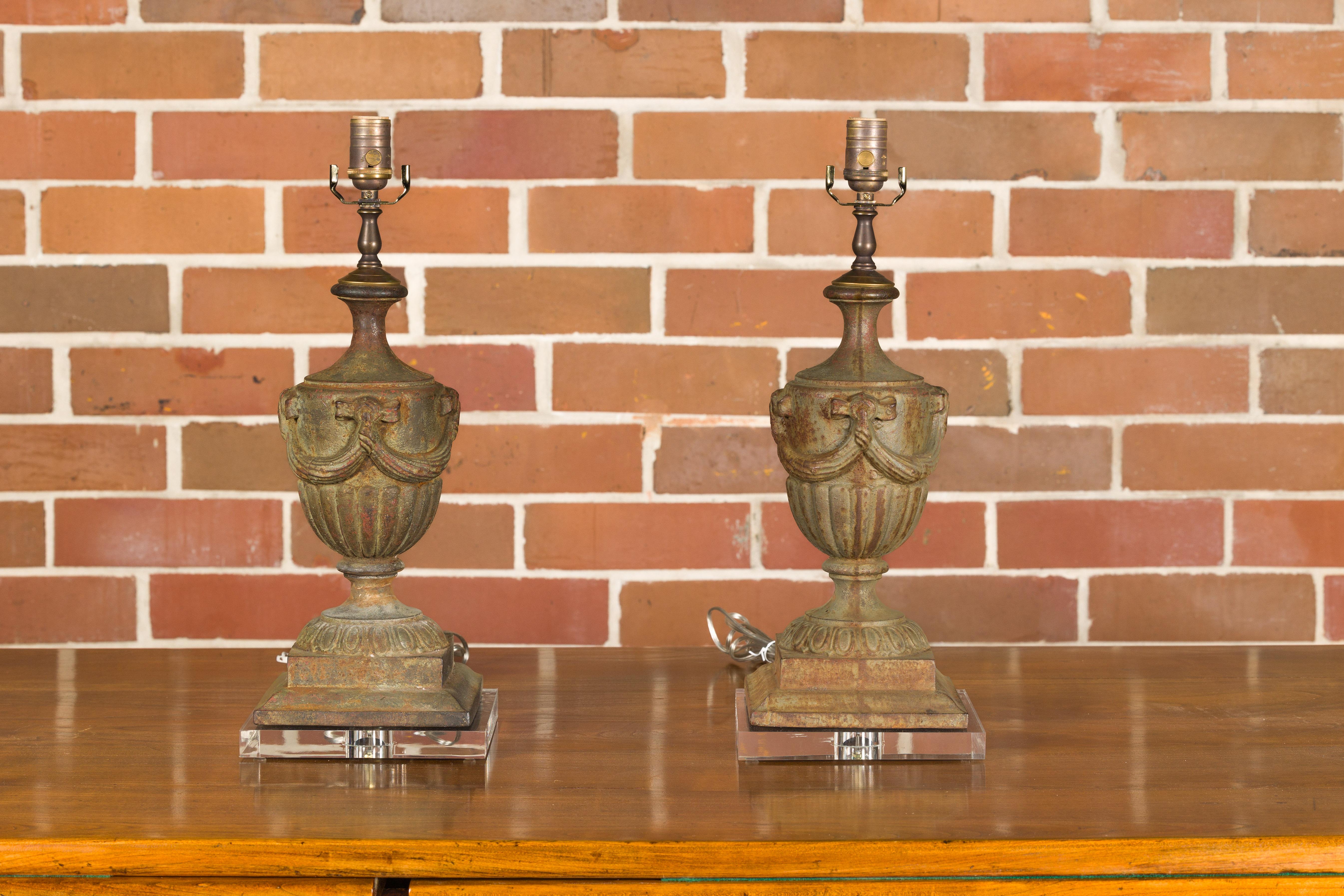 A pair of Neoclassical style iron vase table lamps from the mid 20th century with swag and gadroon motifs, mounted on custom lucite bases. Behold this stunning pair of mid-20th-century Neoclassical-style iron table lamps, a testament to timeless
