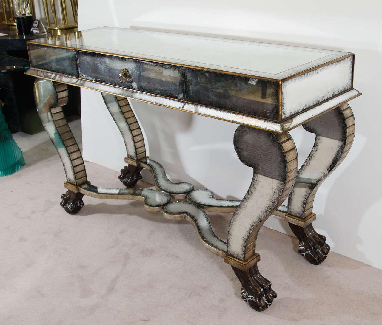Neoclassical style console with central single drawer, covered in aged and distressed mirror panels framed by gilt metal borders. The four feet of the console have lion claws atop ball feet. The piece is in good vintage condition.