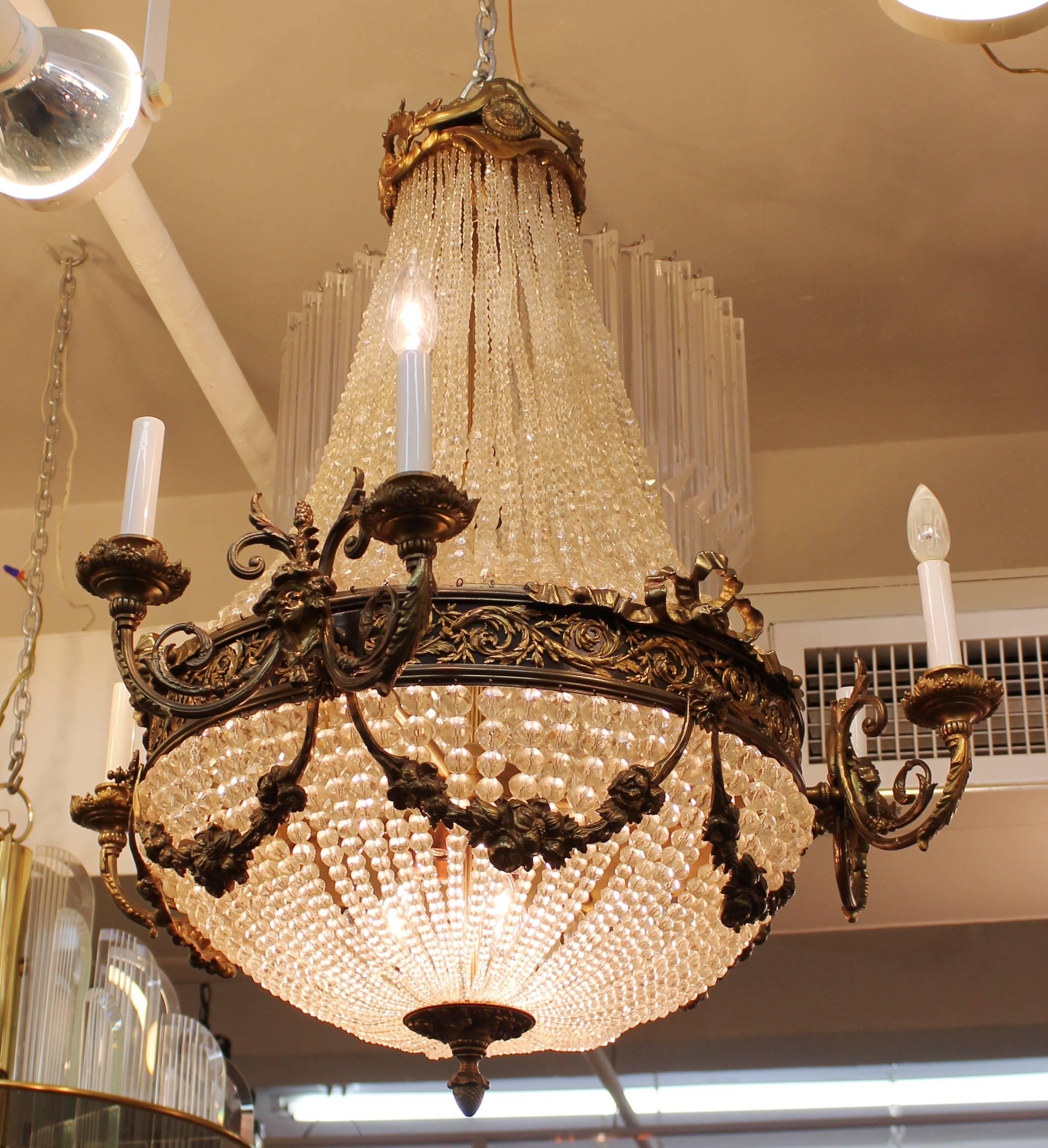 A monumental French neoclassical style chandelier with elaborate bronze decor and cut crystal beads. The sculptural work comprises decorative bronze elements, floral garlands, female allegorical masks and ribbons reminiscent of the style of Louis