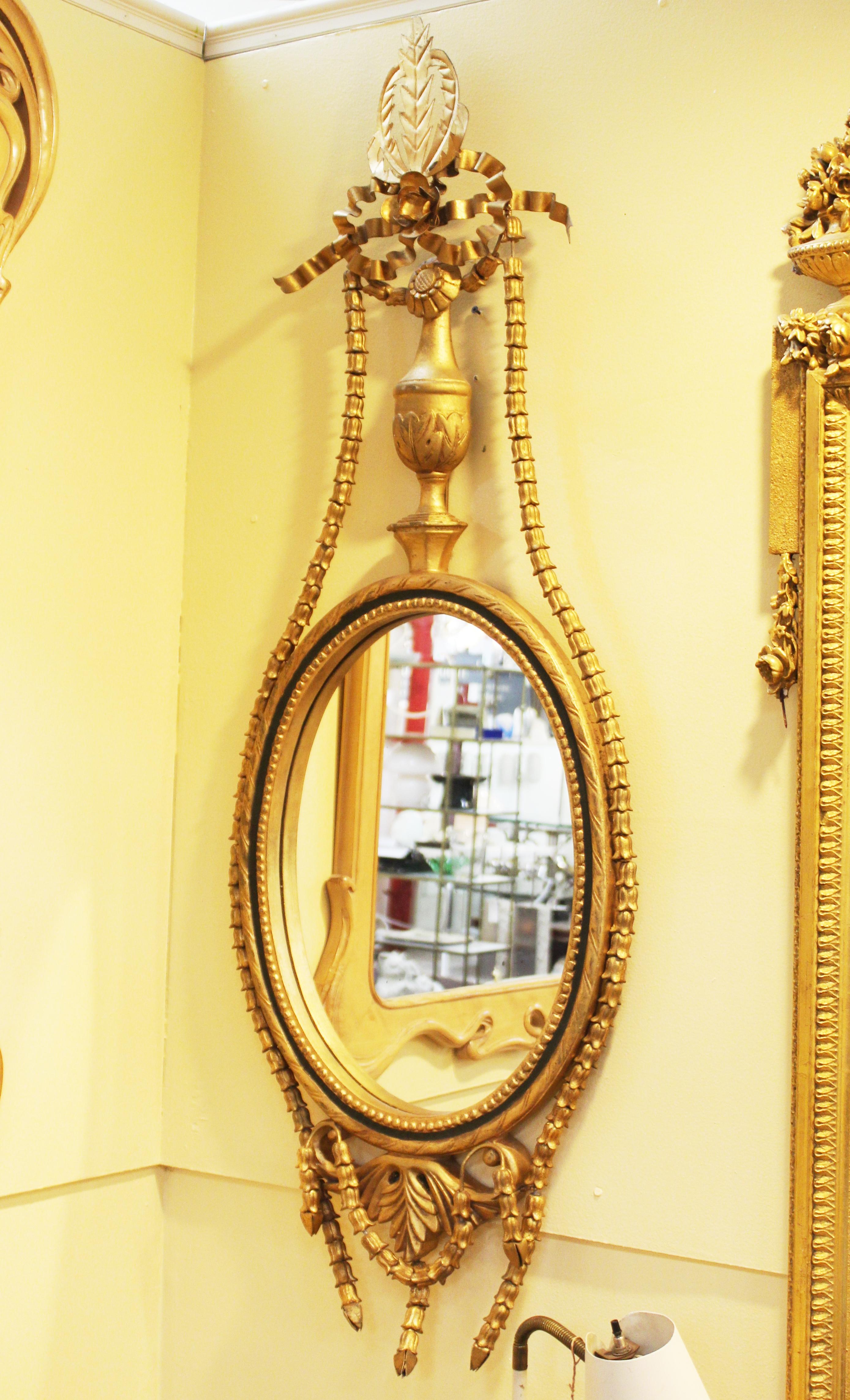 Neoclassical style wall mirror in the shape of a girandole with an oval mirror insert. The piece has floral garlands and ribbons decorating the top and bottom as well as a metal finial. In great vintage condition with age-appropriate wear and use.