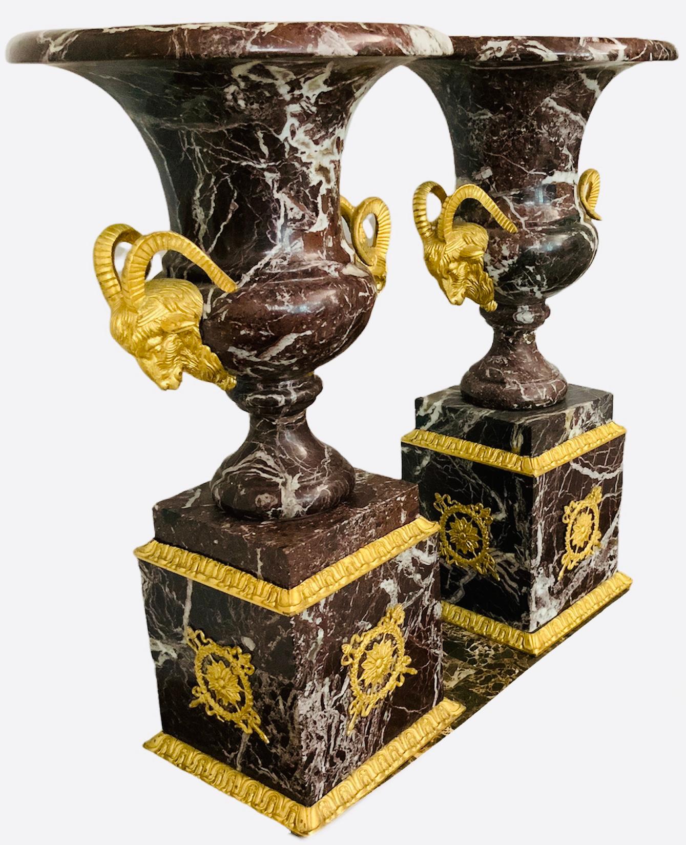 This is a set of brown marble bell shaped urns with gilt bronze ram heads and square marble pedestals. The marble has a bold white veining pattern. The ram heads are placed in the lower part of the body of the urns. The urns have a square shaped
