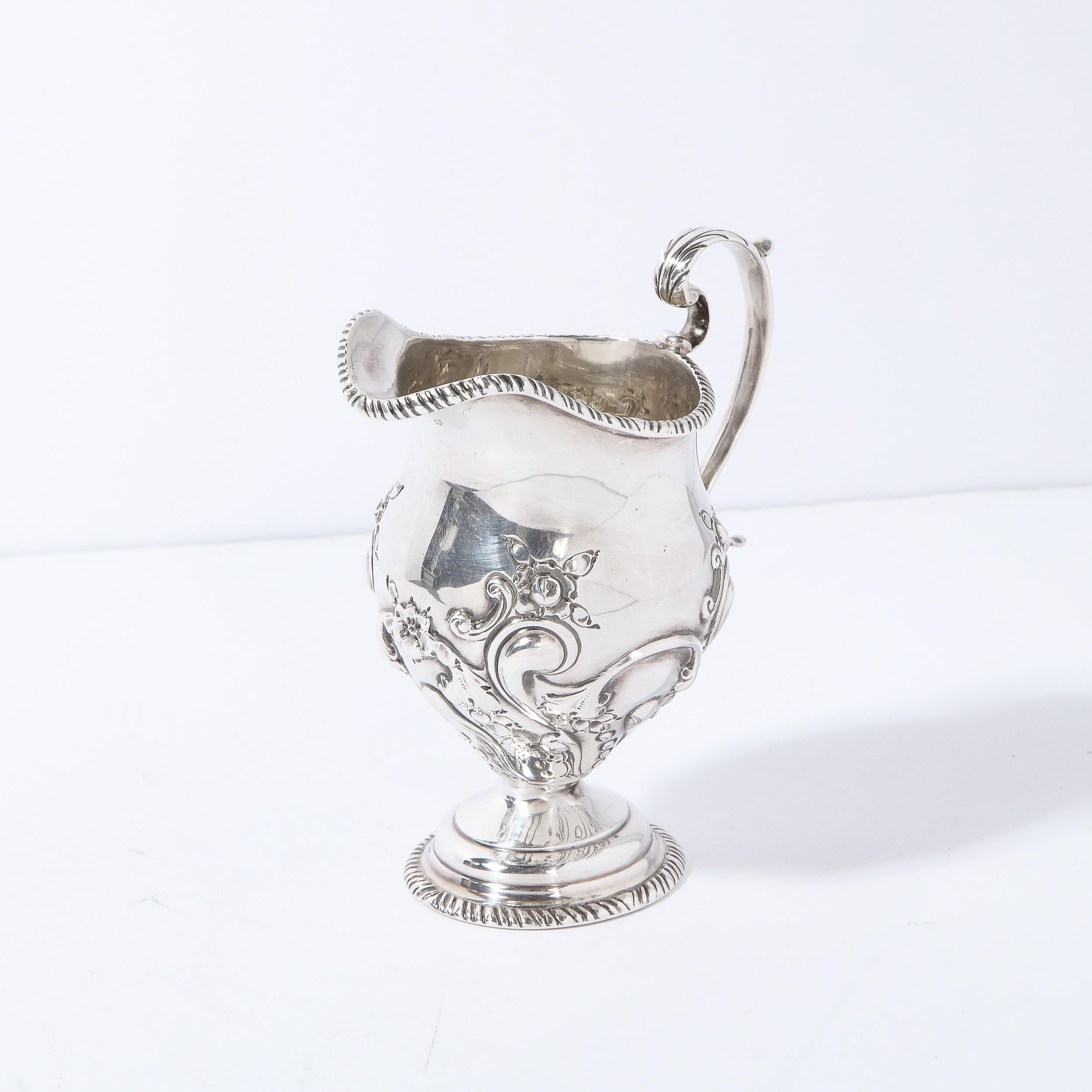 This elegant neoclassical style pitcher was realized by Charles Clement Pilling for Sibray, Hall & Co Ltd in London  circa 1900. It features a channeled base and top; a tiered fillet; a scroll form curvilinear handle; and a rounded body with a