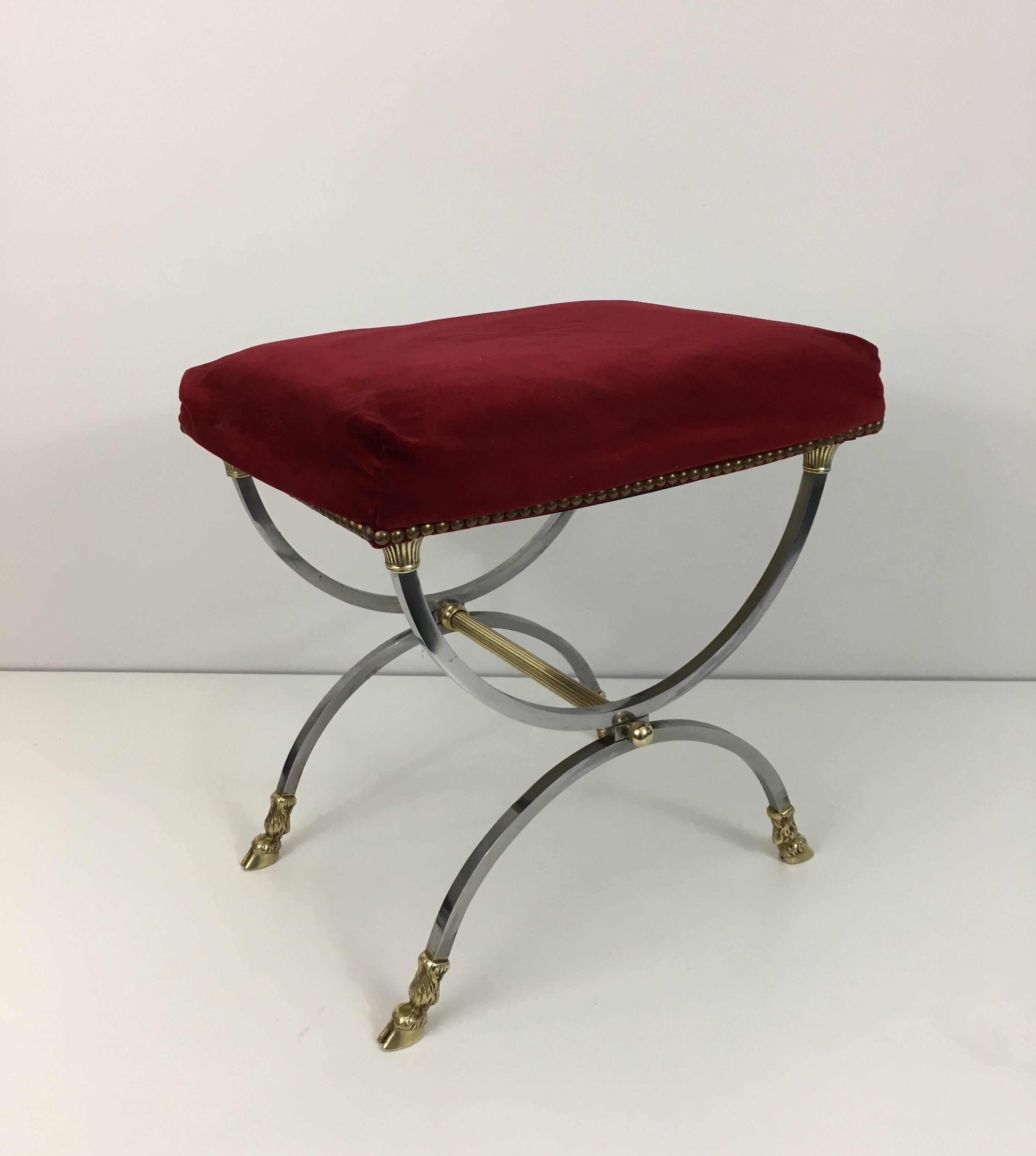 An elegant neoclassical style stool made in brushed steel with a red velvet upholstered seat. The base is comprised of brass hoofed feet and brass fluted stretcher. The seat is resting on bronze capital decorations. French 1940s by Maison