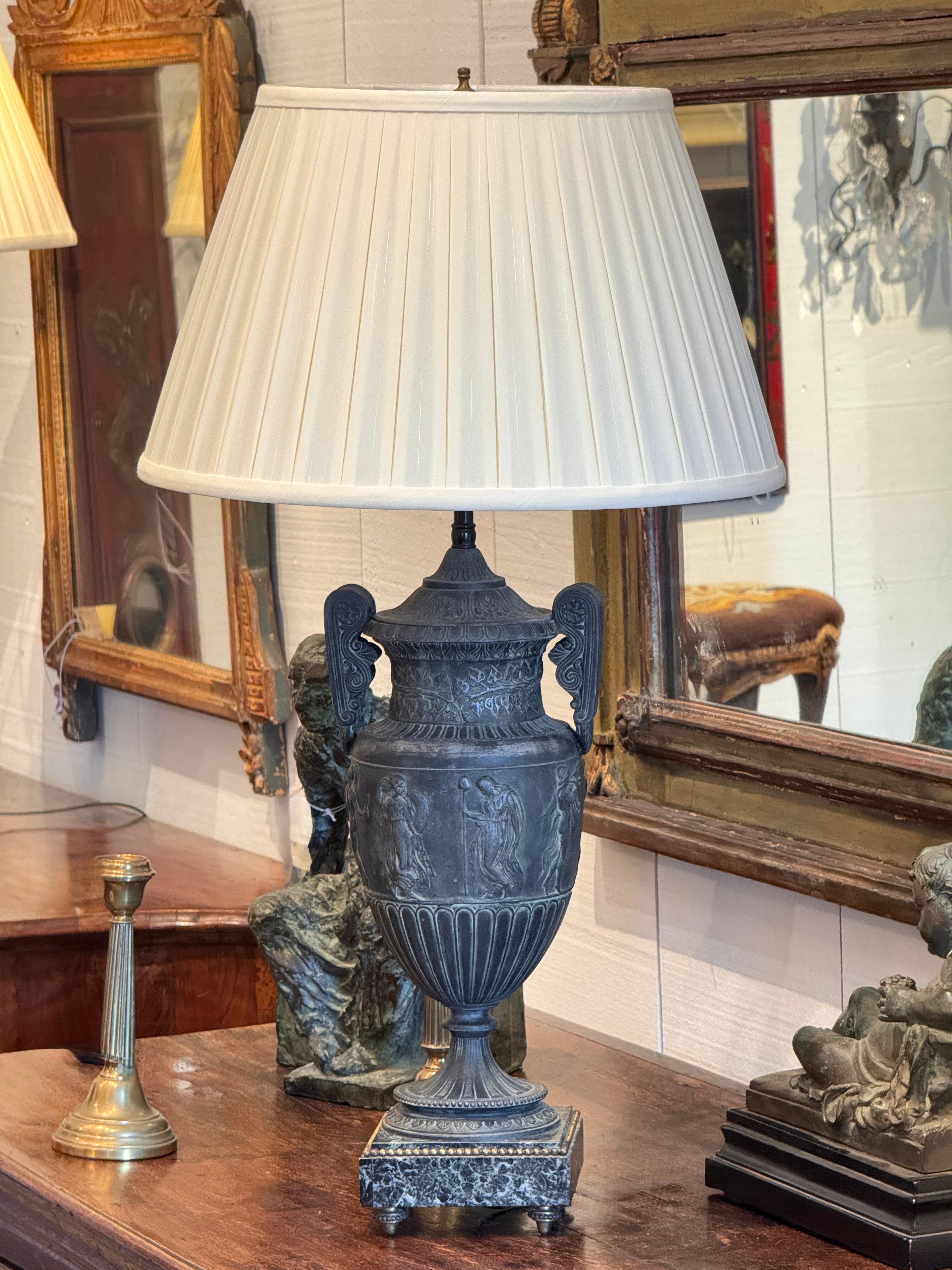 Neoclassical-style table lamp cast in the form of a lidded urn with twin scroll handles and classical figural relief panels. The fluted base rests on a squared plinth with a faux-marble finish. Rich detailing throughout the body and lid reflects the