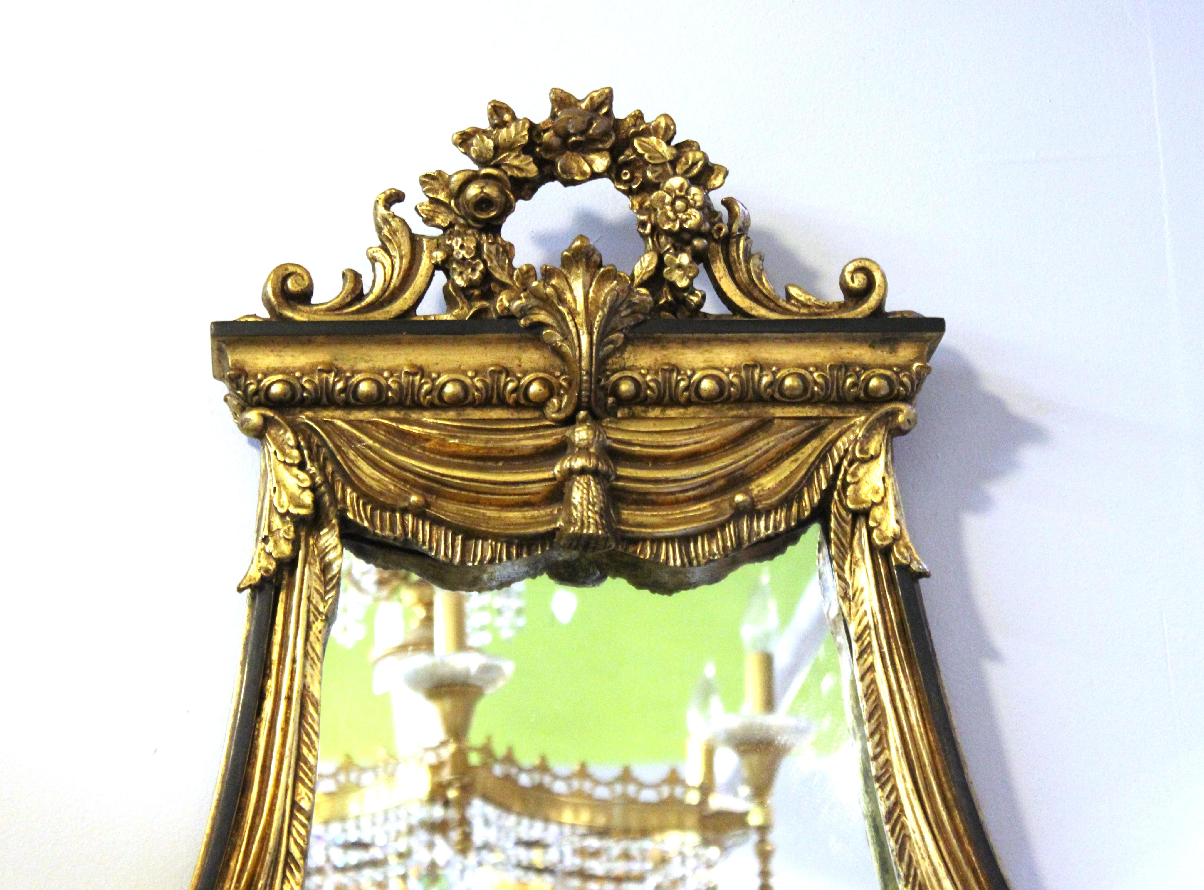 Neoclassical Revival style wall mirror in giltwood frame, with decorative drapery surmounted by a laurel reef and volutes. The piece has a label on the back from 'Decorative Arts Inc. New York' and is in great vintage condition with age-appropriate