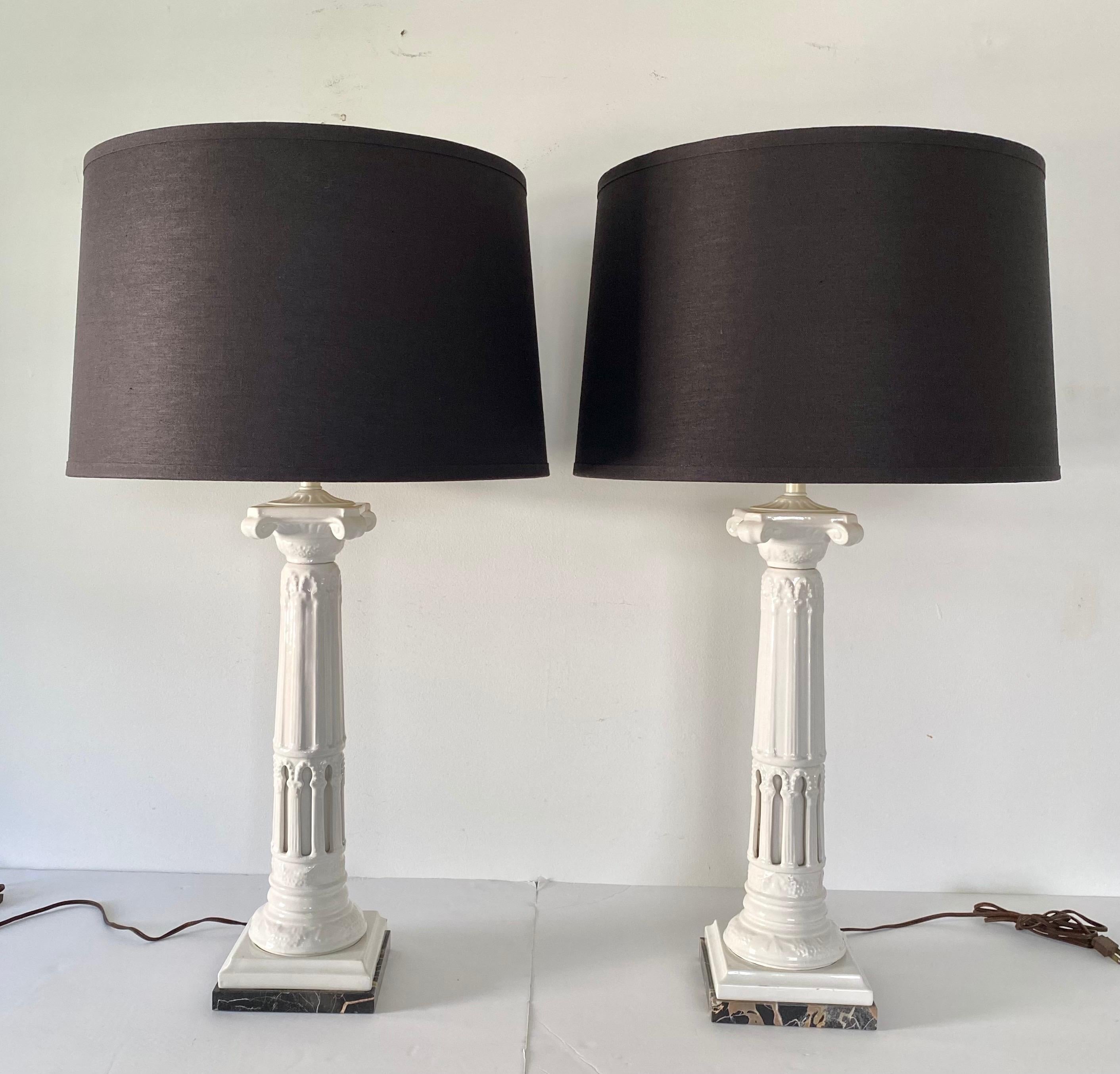 Pair of tall Neoclassical style ceramic fluted column table lamps mounted on black and brown veined marble plinth bases. These sculptural columnar form lamps feature a gloss white glaze with draped garland reliefs and dimensional cut-outs. Lamp