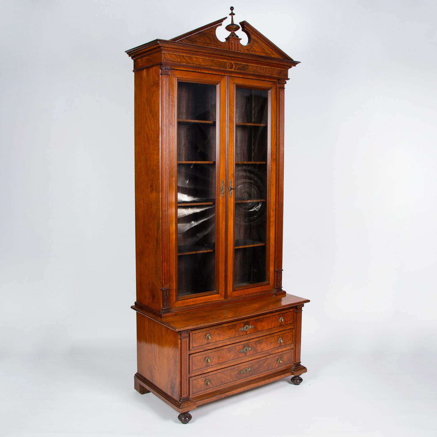A neoclassical walnut two-door bookcase, with broken pediment, with two glazed doors flanked by fluted pilasters, over three drawers, on bun feet. 

Walnut, burr walnut and ebony stringing.

Brass handles and escutcheons.

interior with four