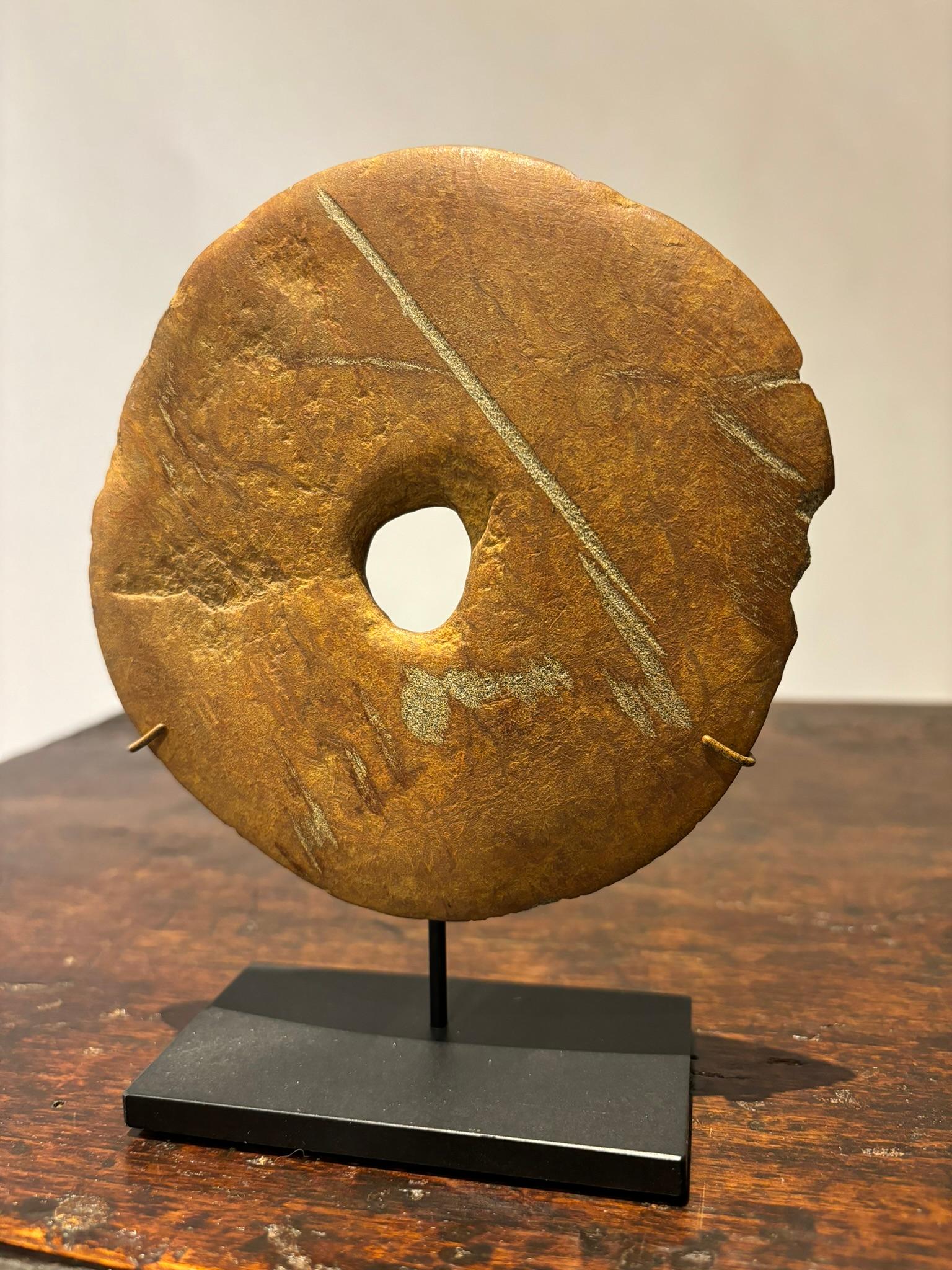 Disco neolítico de piedra tallada a mano con un agujero en el centro, montado sobre una base moderna de acero. Esta evocadora pieza antigua tiene una presencia real que desmiente su tamaño. Maravilloso color con bonito estriado de contraste. Esta