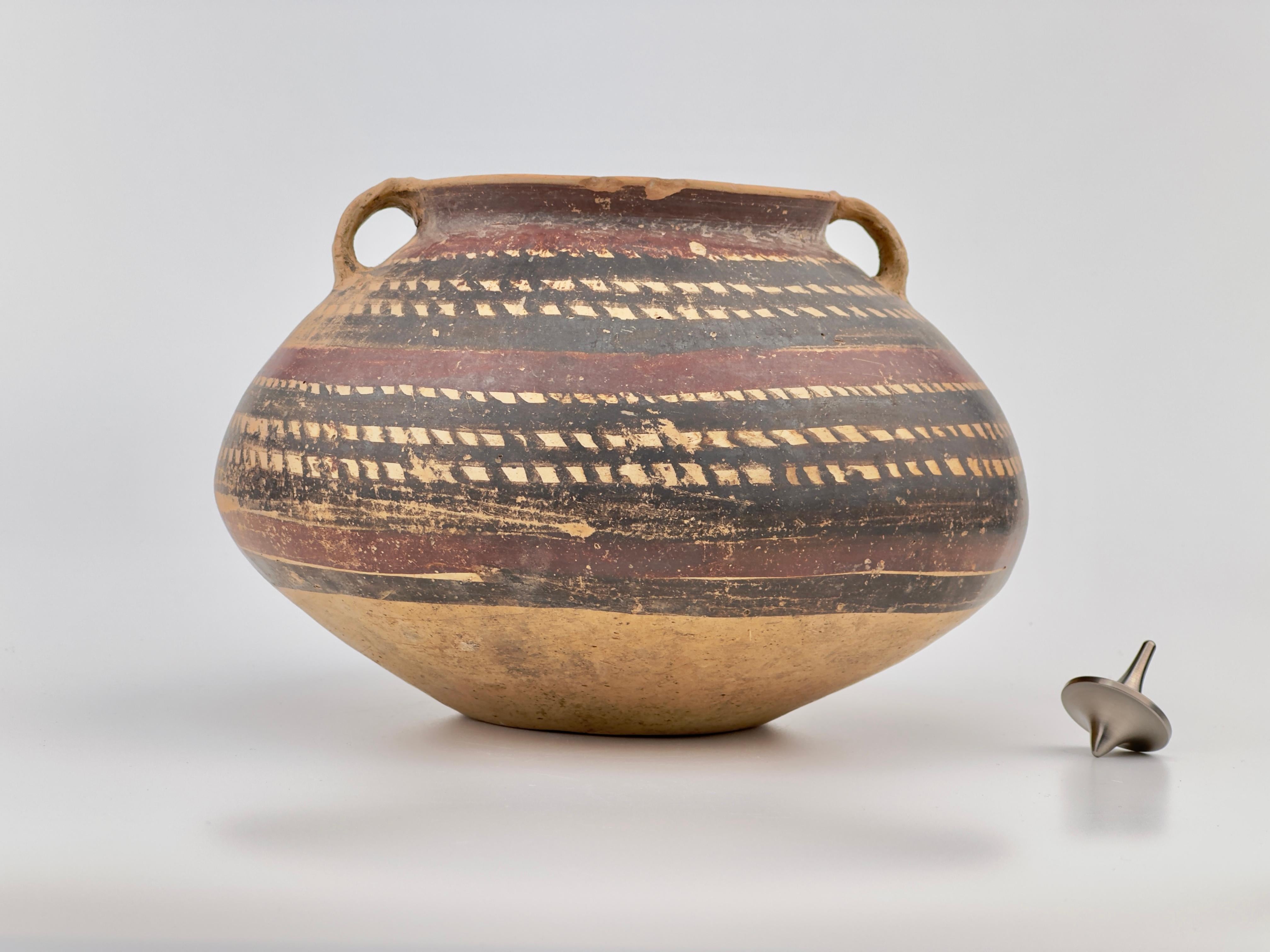 This pottery has a bulbous body with two small, protruding handles near its widest part. The pottery features horizontal bands of patterns, which include a sequence of geometric shapes and lines. The patterns are rendered in a dark pigment, possibly