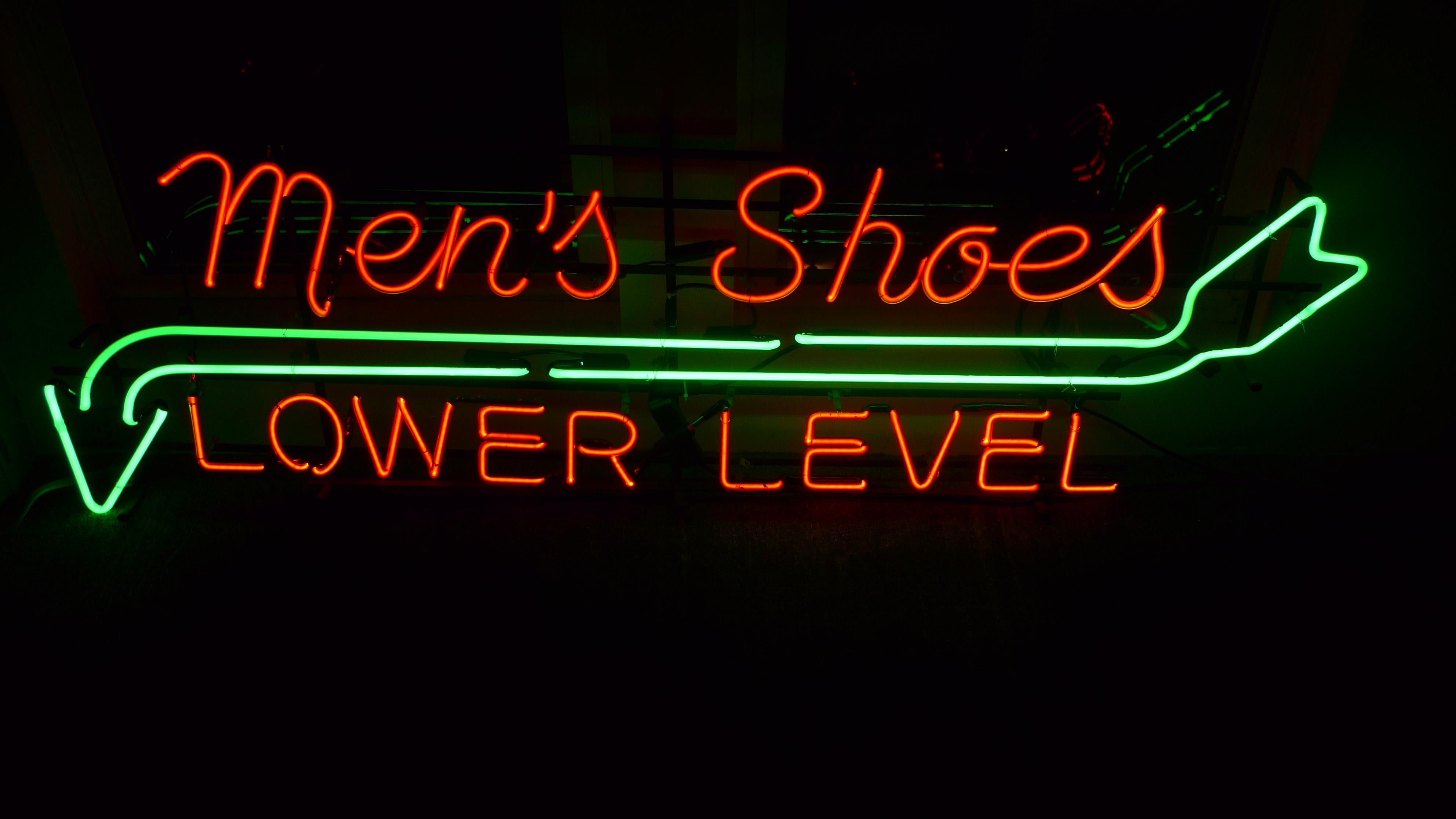 Neon signage from a midwestern department store directing customers to: Men's shoes, Lower Level, circa 1930s. Strikingly colorful in red-orange and green neon designed around a symbolic arrow splitting cursive and sans serif type. Haute design at