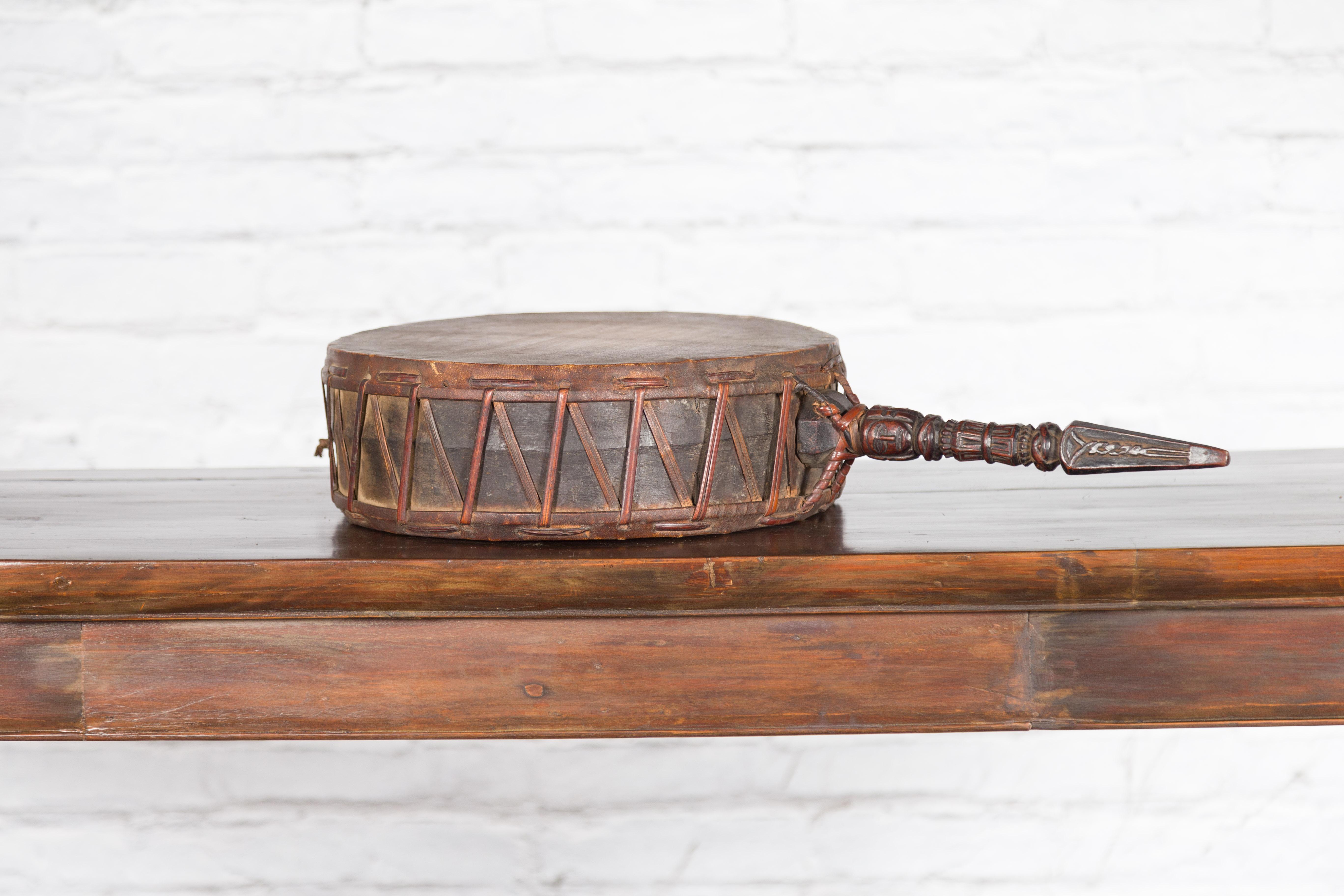 Ancien tambour à main népalais en cuir du XIXe siècle, avec un manche en bois sculpté à la main. Créé au Népal au cours du XIXe siècle, ce tambour à main se compose d'une peau circulaire en cuir à l'aspect joliment usé, reliée sur le côté à une