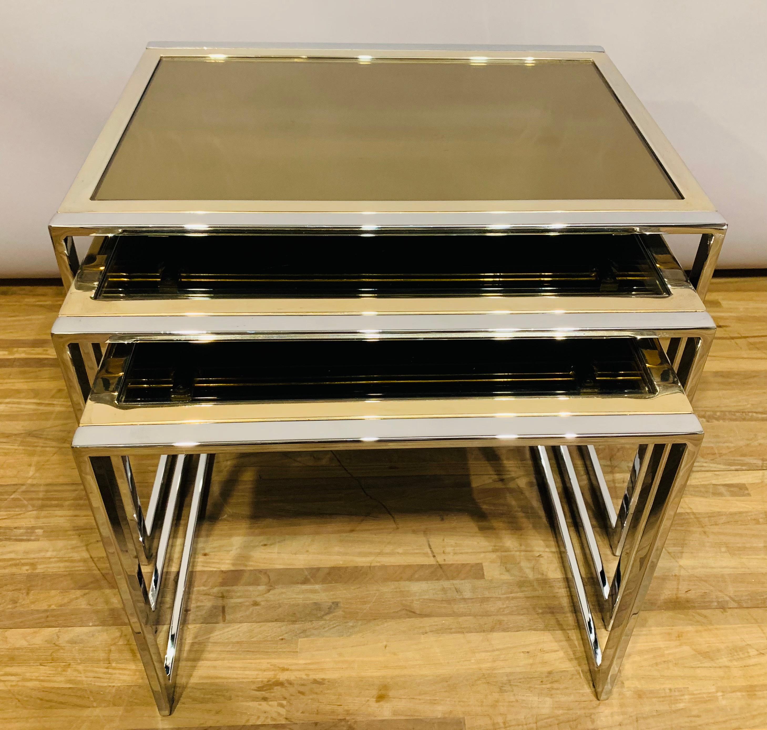 A set of 3 Belgo Chrom nesting tables. The frames are constructed from solid chromed metal with gold chromed trim surrounding the smoked glass mirrored table tops. The mirrored table tops sit within the frame. There are some scratches to the glass