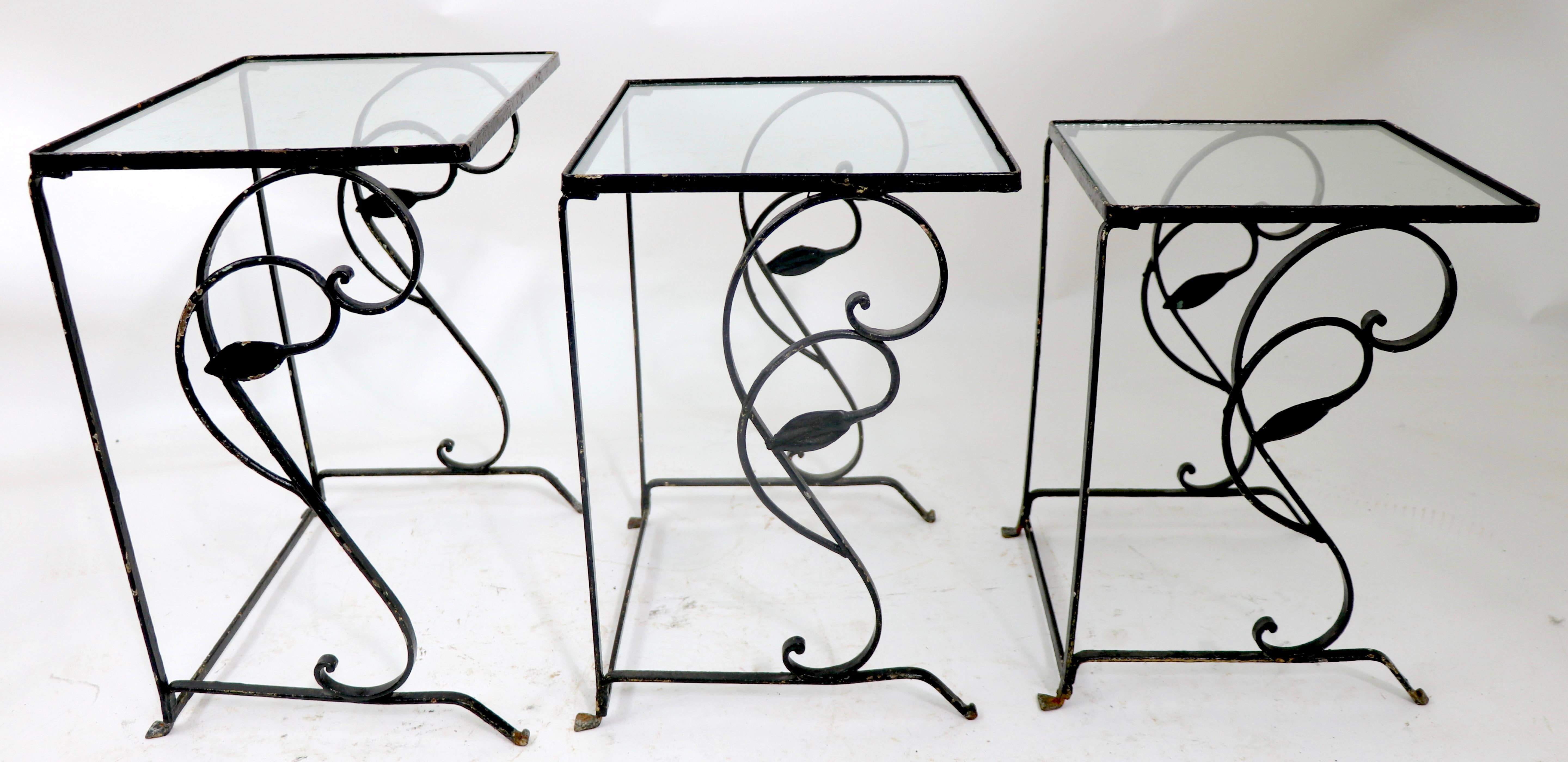 Chic three piece set of graduated nesting tables having decorative wrought iron frames and glass insert tops. The tables are structurally sound and sturdy, free of damage, or repairs. The tables are currently in later, but not new , black paint