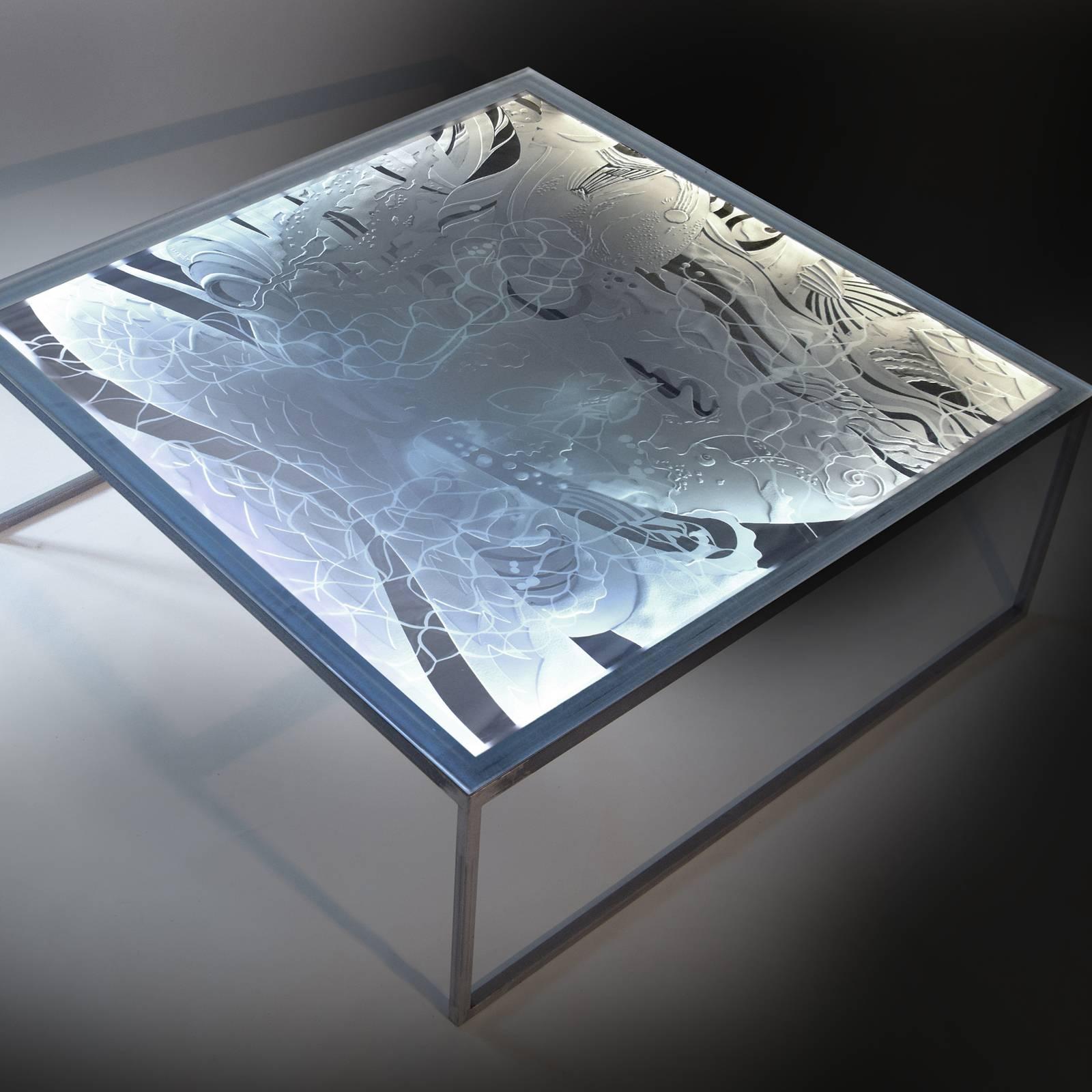 An alluring dance of light and form, this glass table stands out for its originality and fanciful allure. The glass tabletop boasts a marine design in bas-relief achieved exclusively with hand engraving techniques. It is supported by an iron