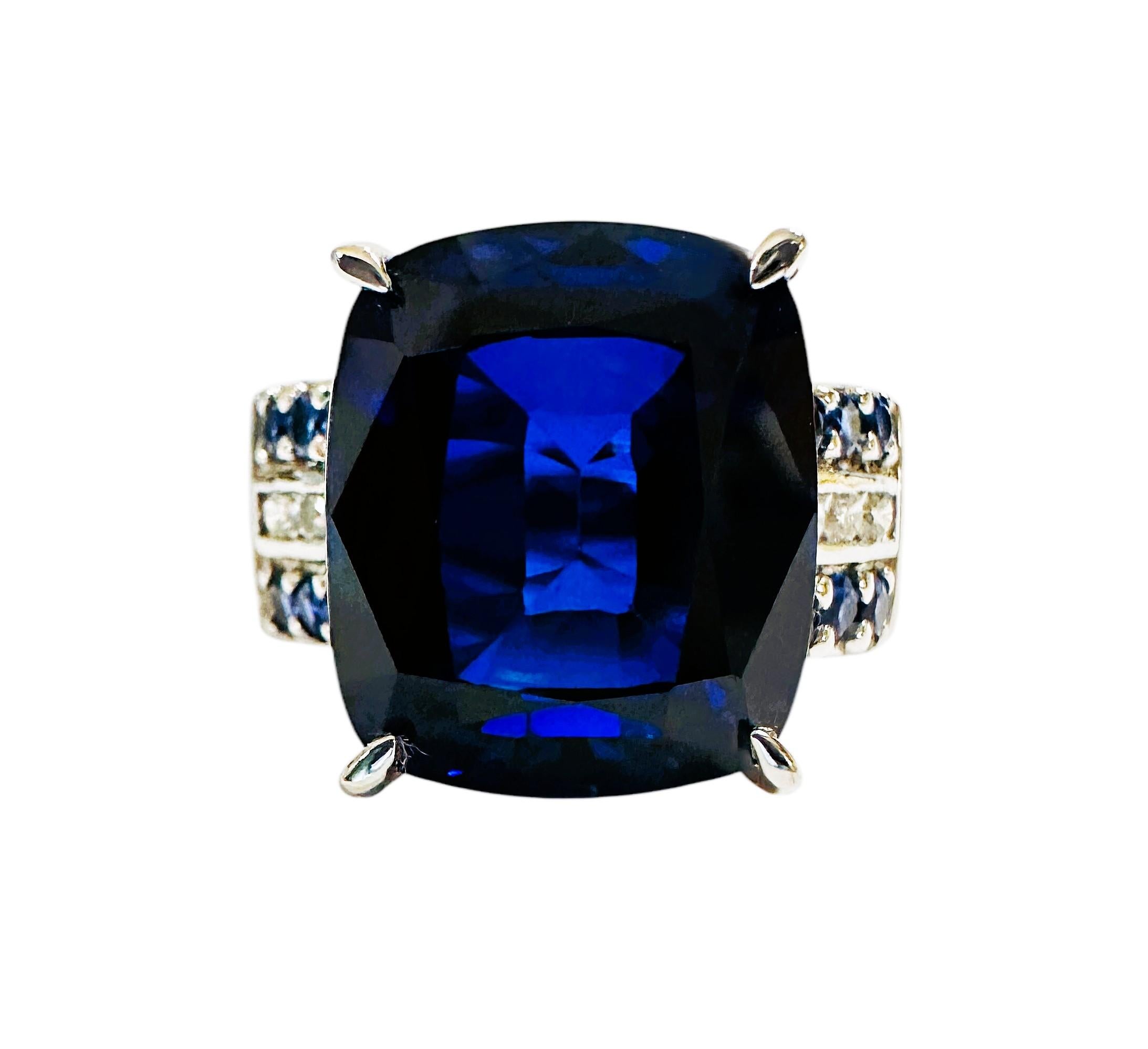 La piedra procede de Sri Lanka y está bellamente tallada.  Es un zafiro azul y tiene 13,9 quilates.  Es una piedra talla cojín de fantasía, mide 14,3 x 12,8 mm y está flanqueada por 2 filas de piedras de zafiro azul y blanco talla diamante a cada