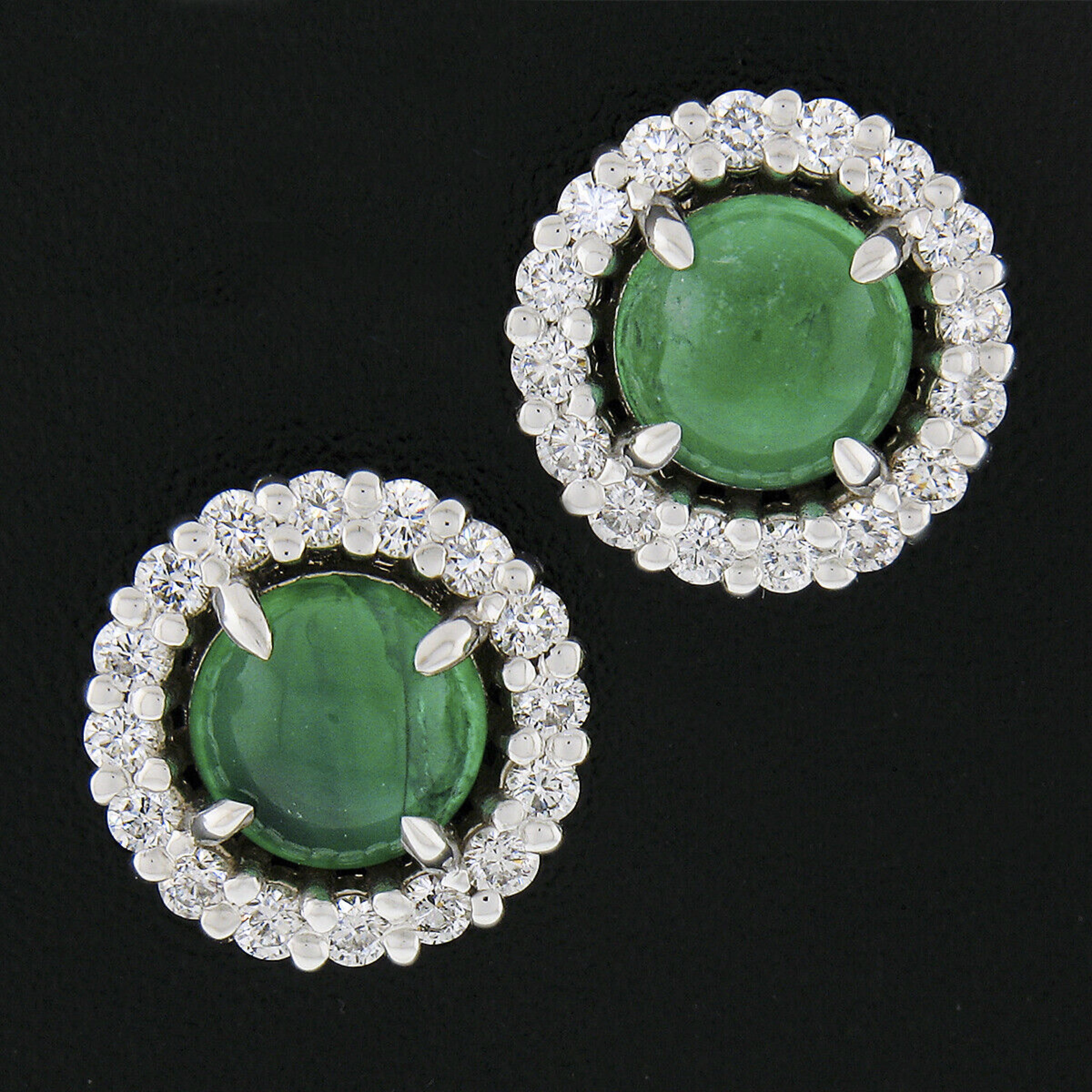 This gorgeous pair of emerald and diamond halo stud earrings is newly crafted in solid 18k white gold. Each earring features a gorgeous round cabochon cut natural emerald stone neatly prong set at its center, and are well matched displaying the most