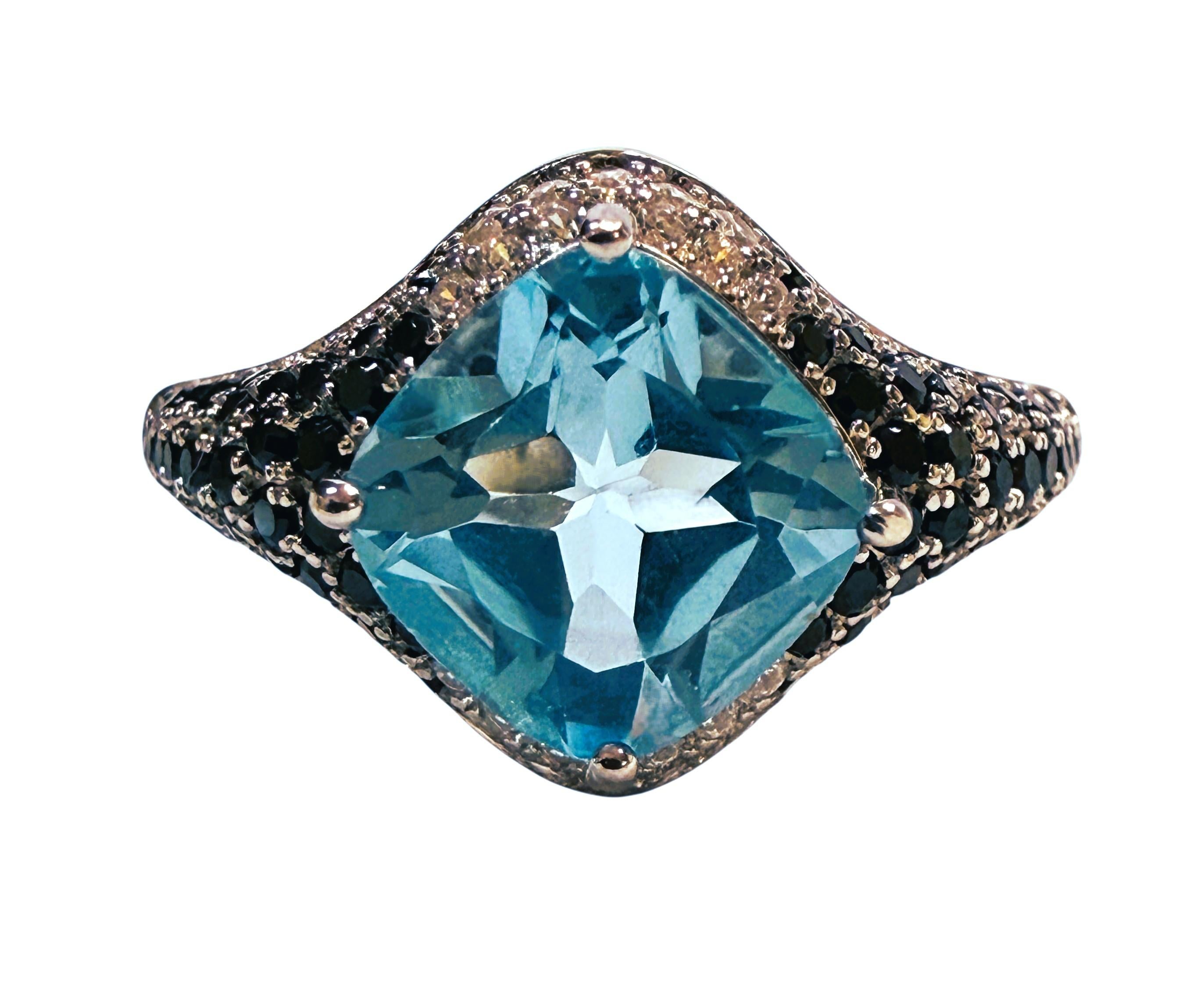 Es un anillo muy singular y bonito. La piedra es un topacio azul tallado en cojín y mide 2,04 quilates.  Mide 8 x 8 mm.  Está rodeada en parte por espinelas negras talladas con diamantes, que también se extienden por la banda.  También hay zafiros